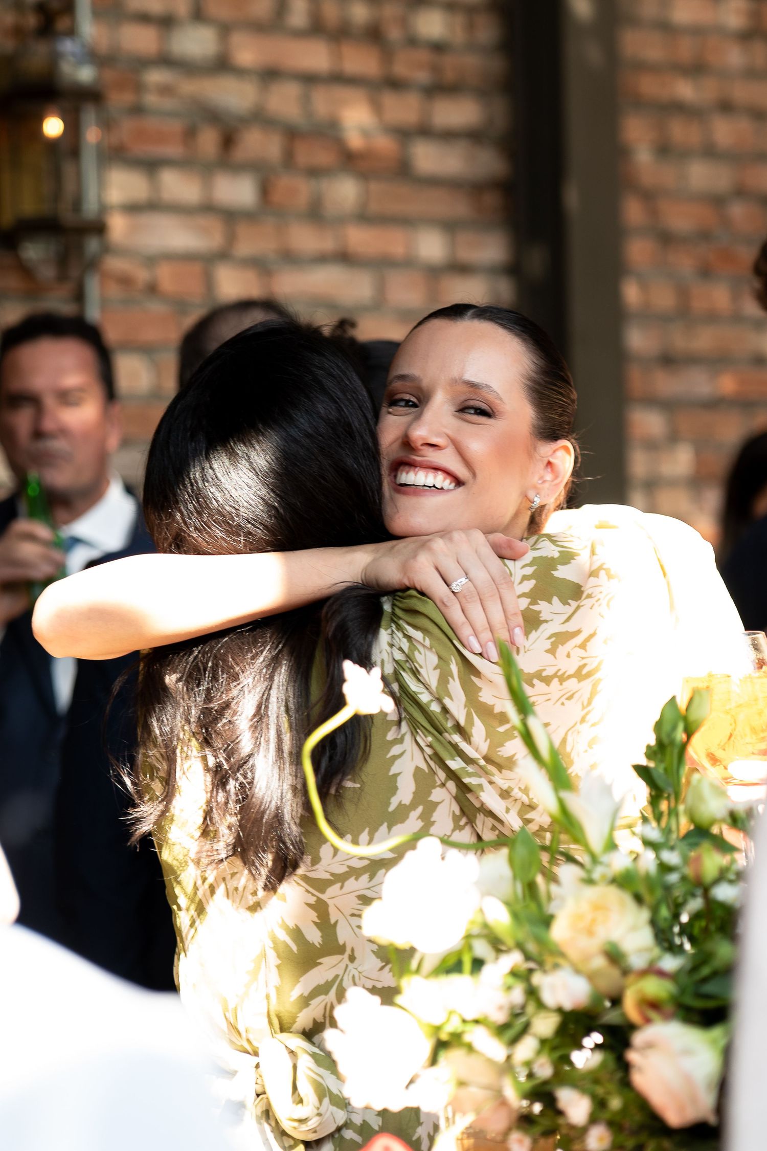 Two women share a warm hug at a wedding, one in a floral dress smiling, with flowers and brick wall in background.
