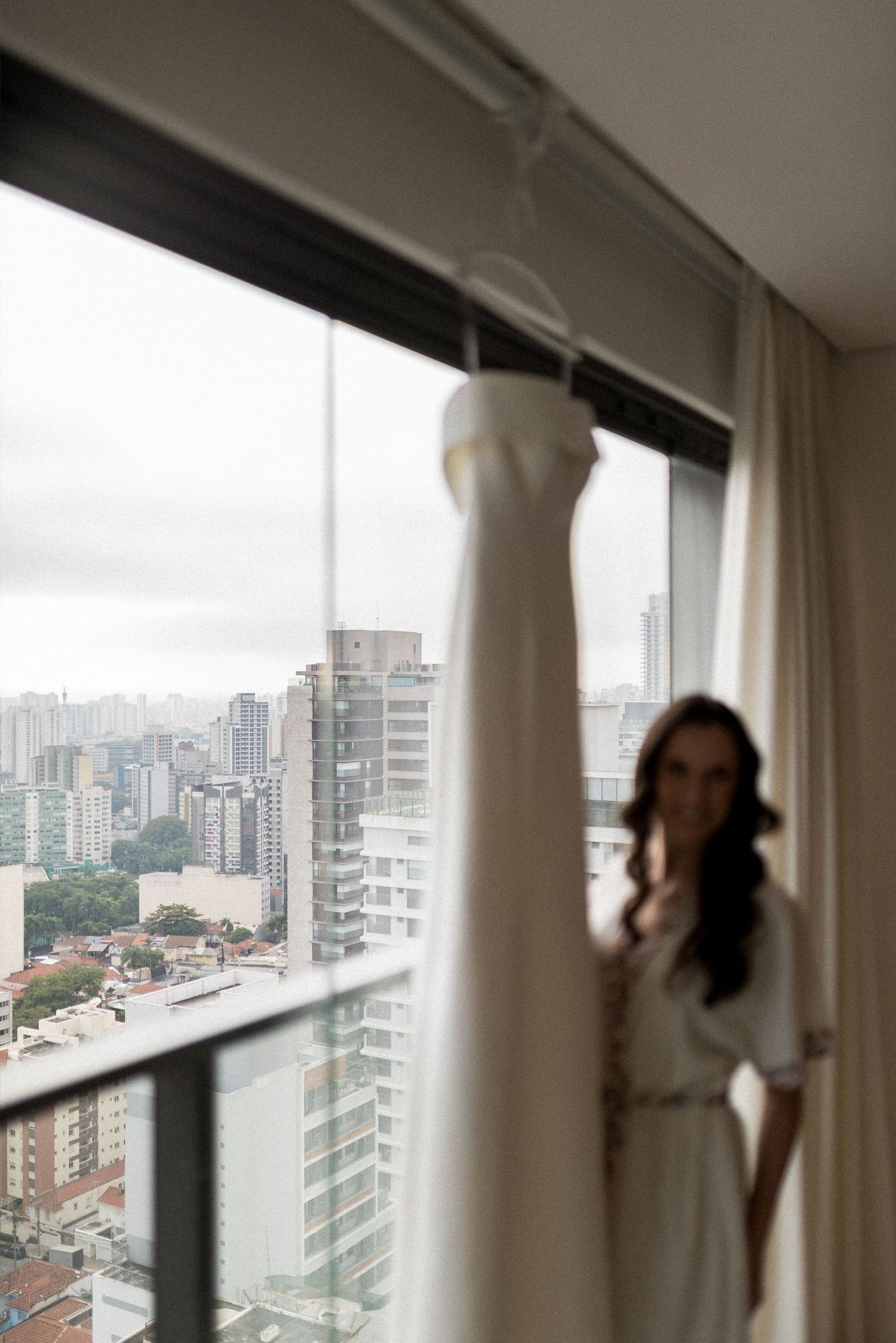 A bride in a white robe stands by a high-rise window with her wedding dress hanging in the foreground, city skyline behind.