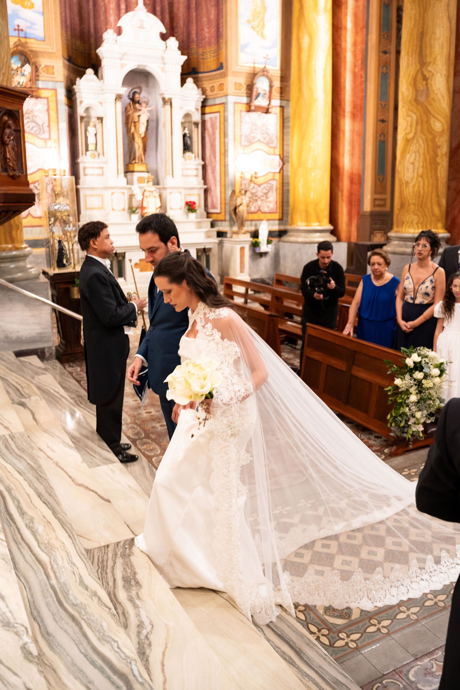 Bride in lace wedding gown and veil walks down church aisle with groom during elegant Catholic wedding ceremony.