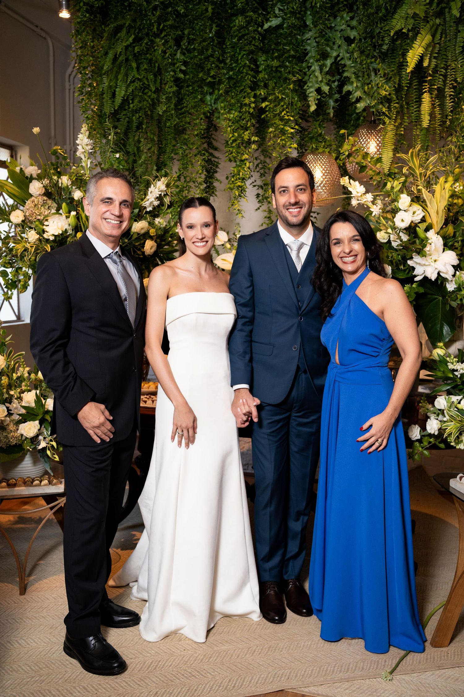Wedding party of four posing before lush floral backdrop, bride in white gown, groom in navy suit, guests in blue dress.