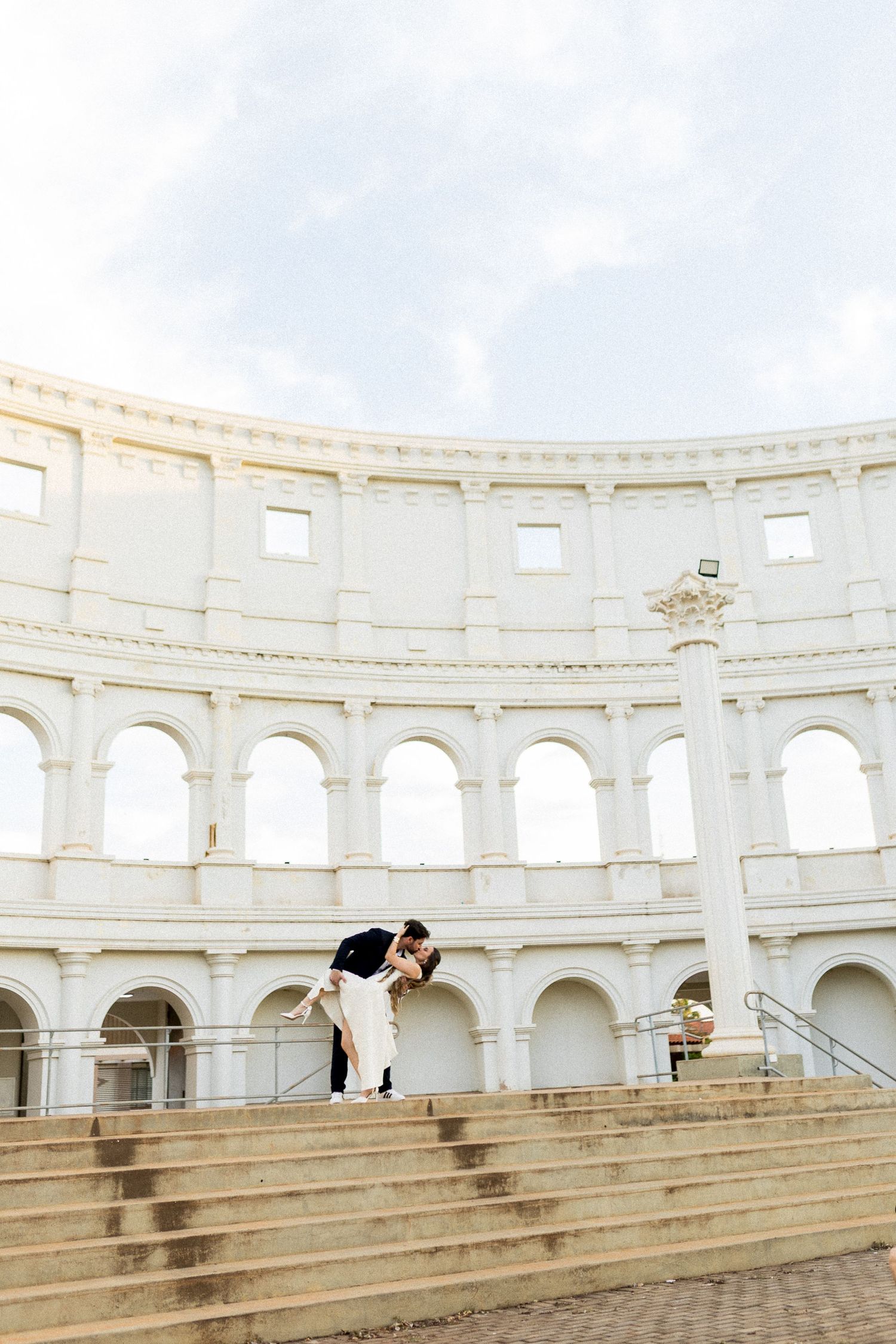 Couple sharing a romantic dip kiss on steps of a white Roman colosseum-inspired wedding venue.