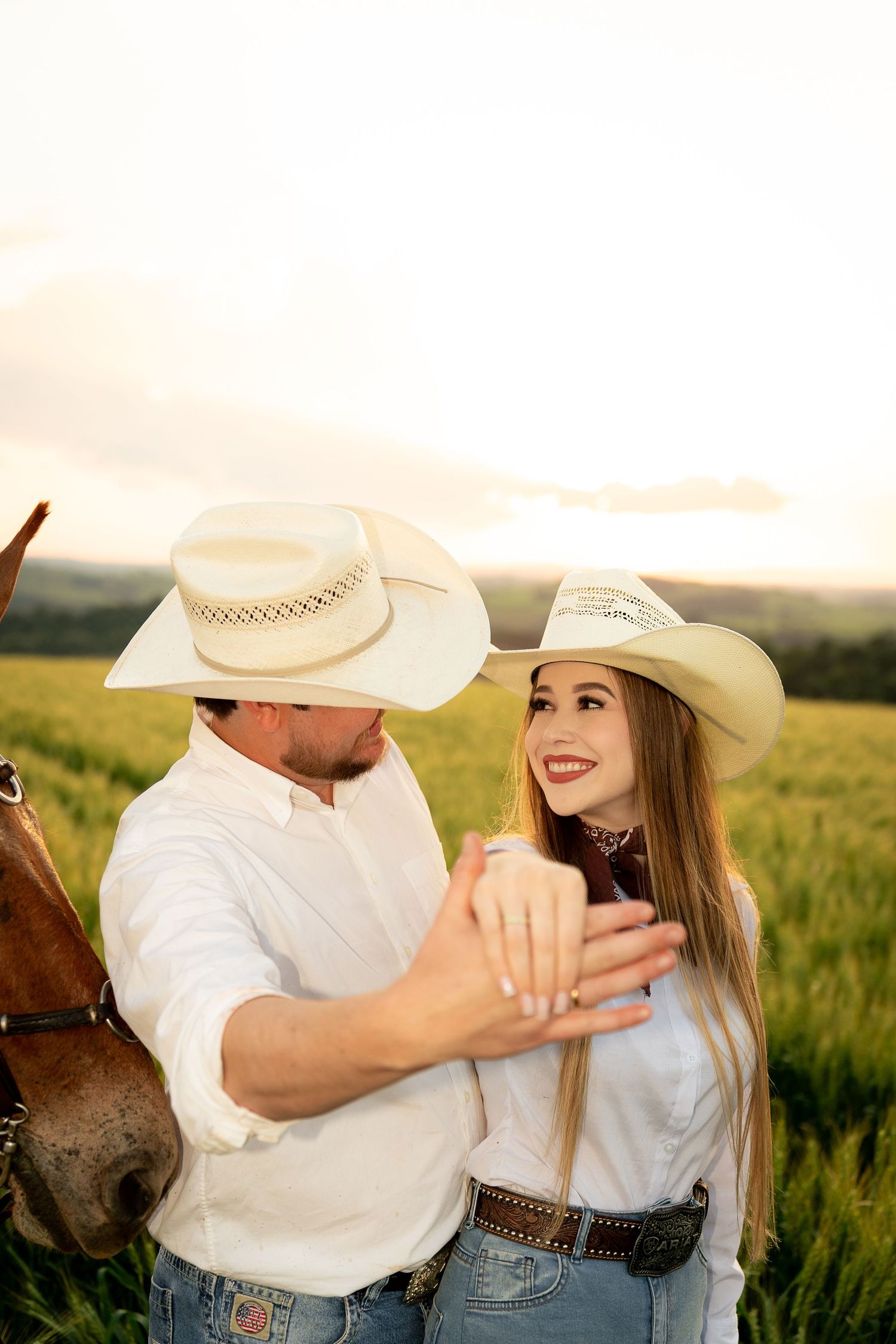 A couple in matching white cowboy hats and shirts embrace in a green field at sunset near a horse.