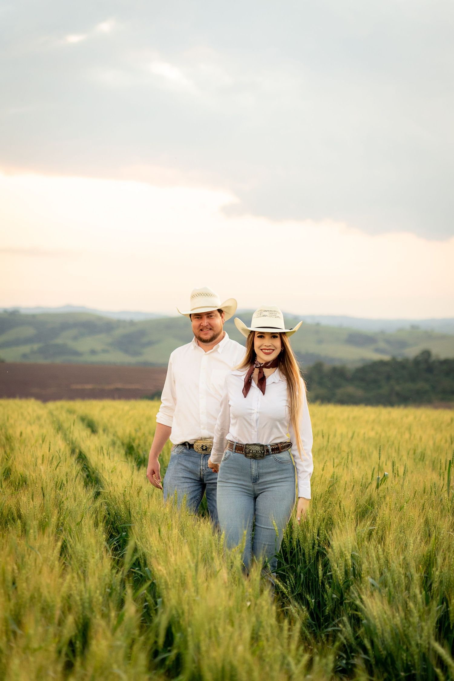 A couple in white shirts and cowboy hats walks through a lush green wheat field at sunset.