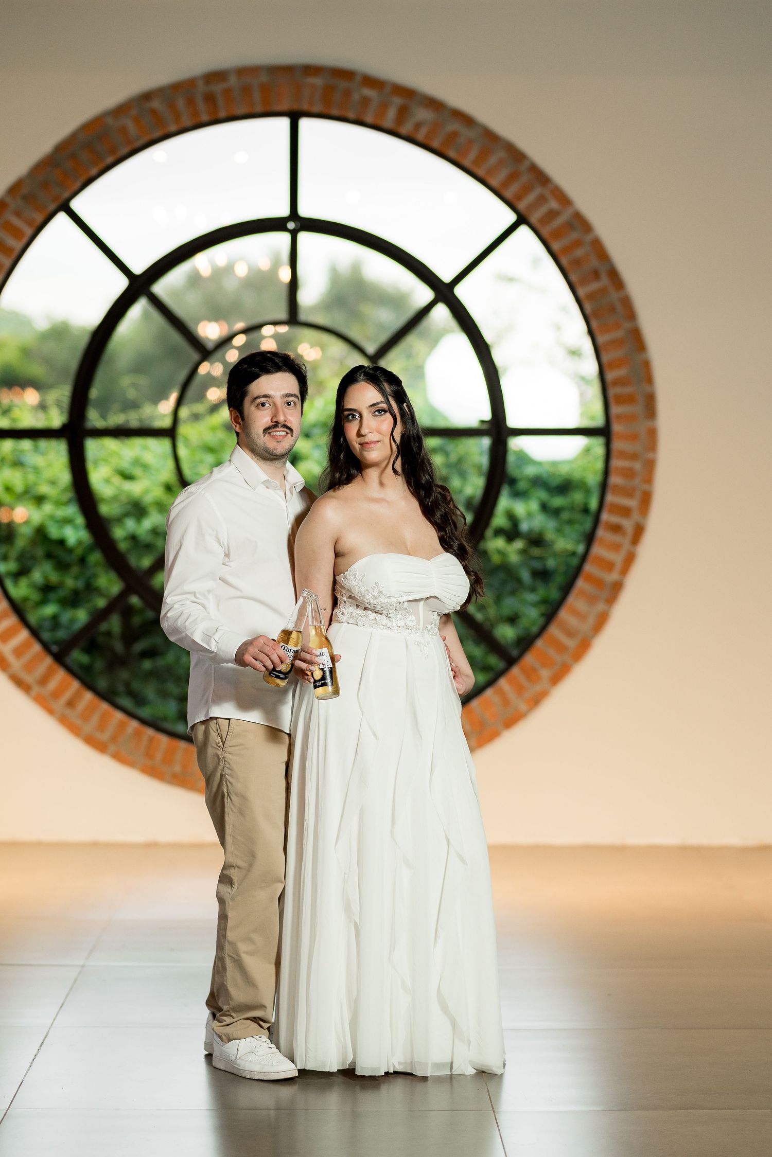 Couple in wedding attire poses before a circular brick window with lush greenery, holding champagne bottles.