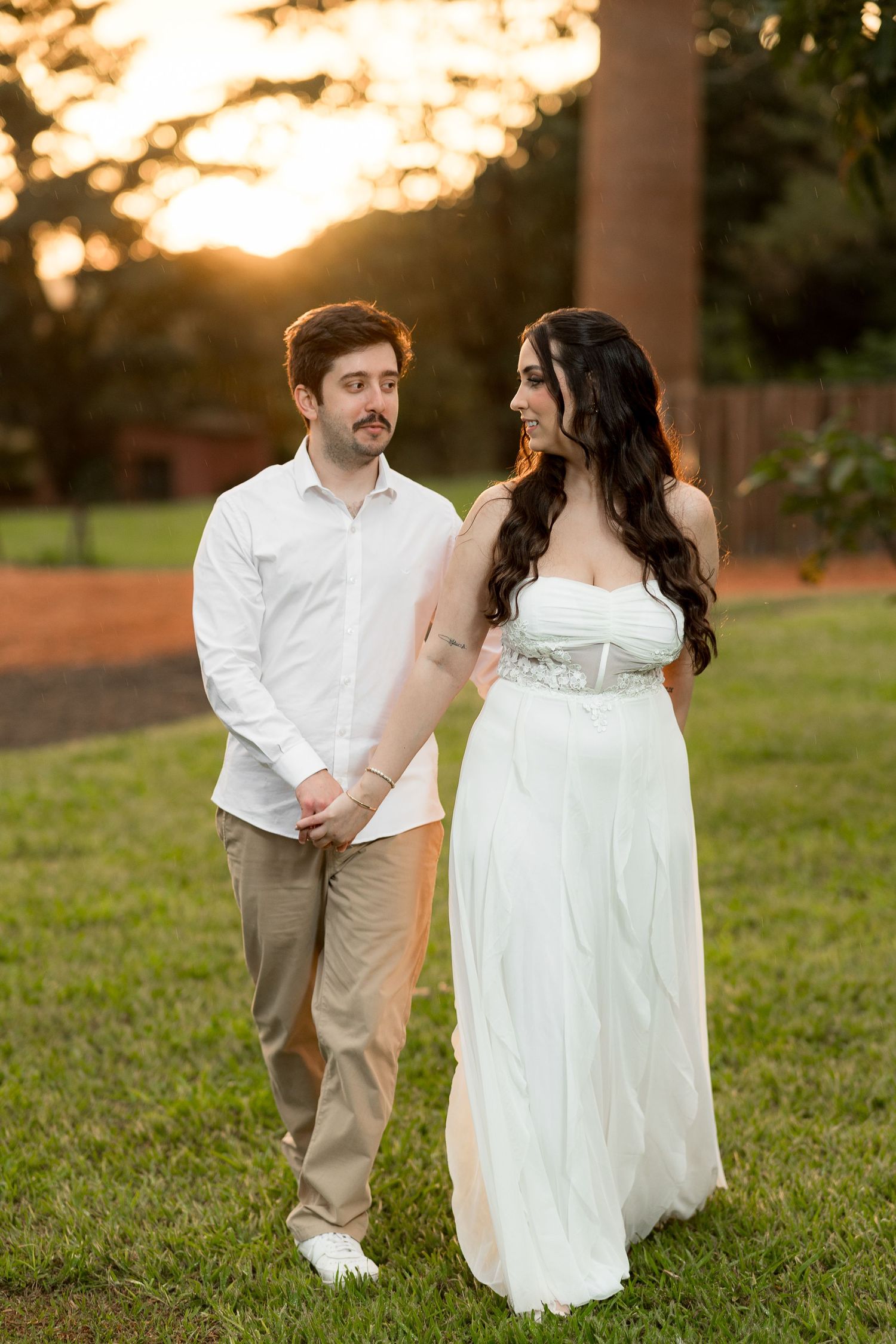 Couple in white attire holding hands at golden hour sunset in a lush green park setting.