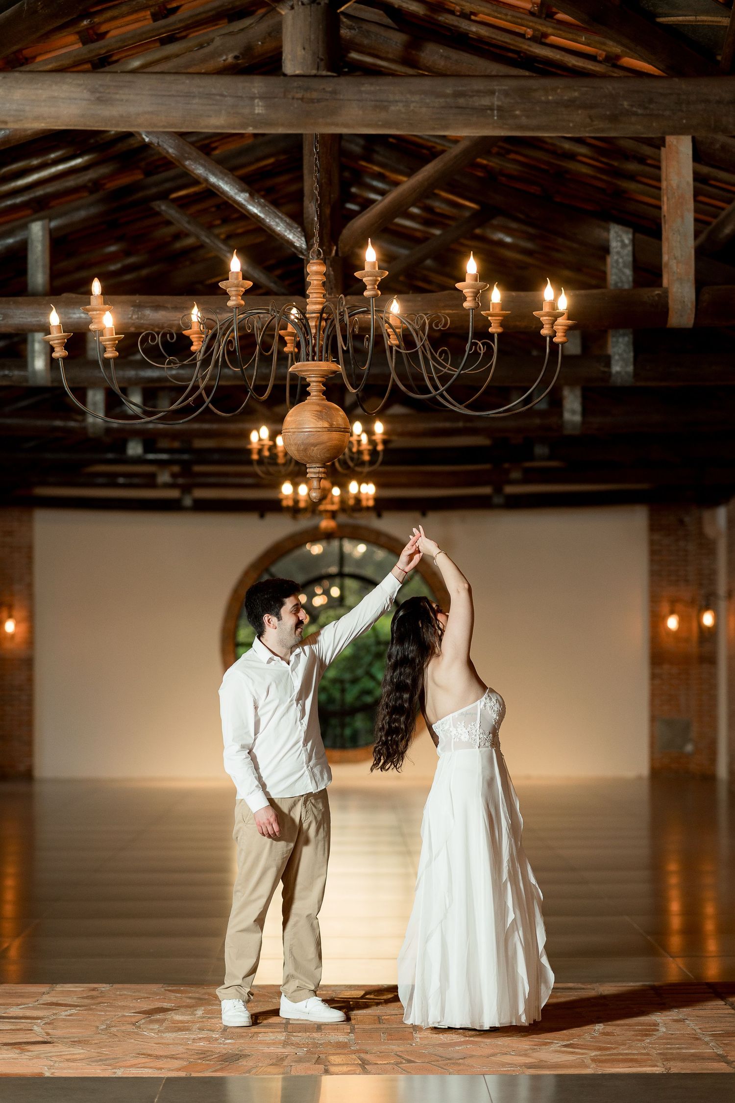 Couple dancing under a rustic chandelier in an elegant venue, bride in white gown being twirled by groom.