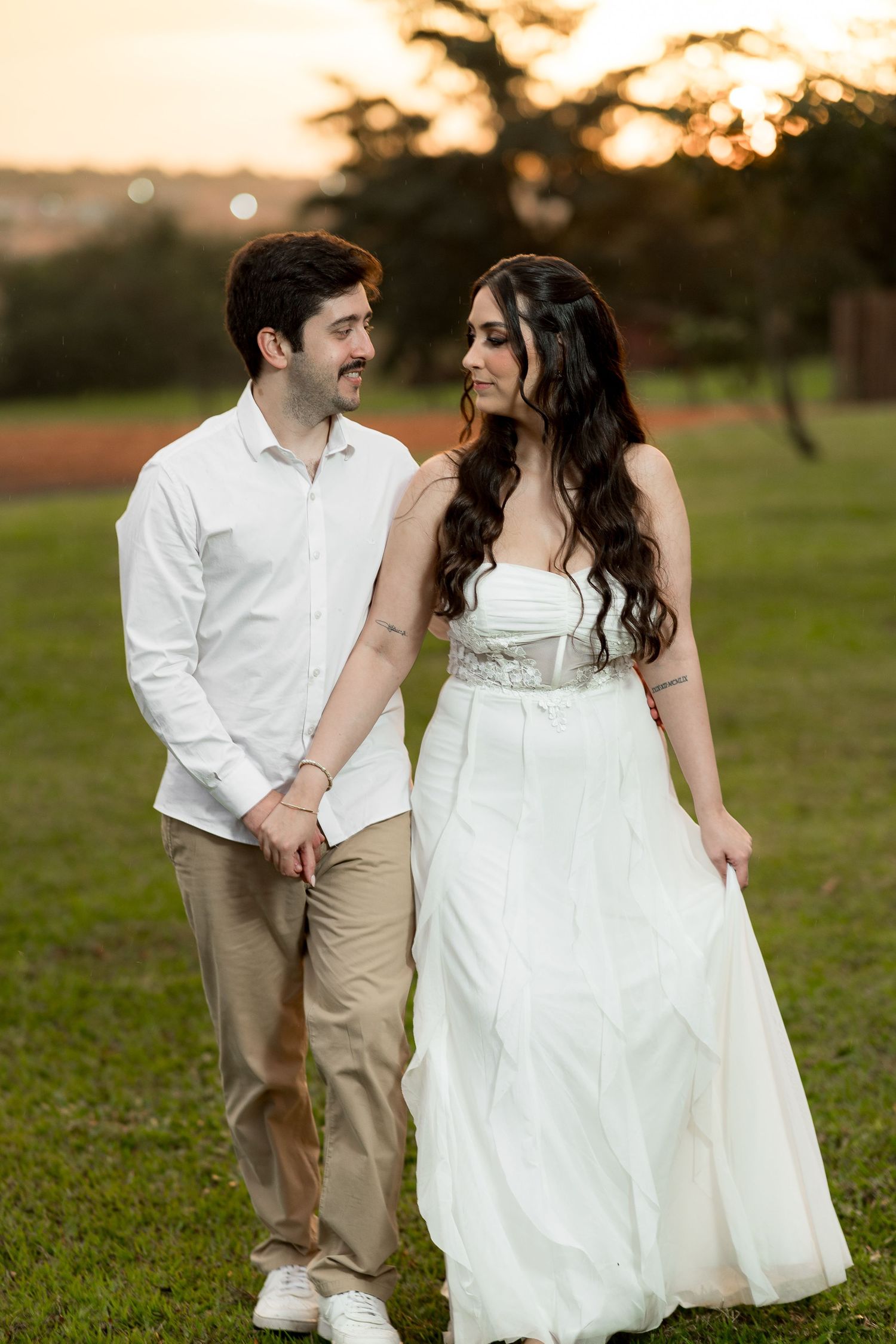 Couple in white attire walking hand-in-hand on green grass at sunset, sharing a romantic glance.