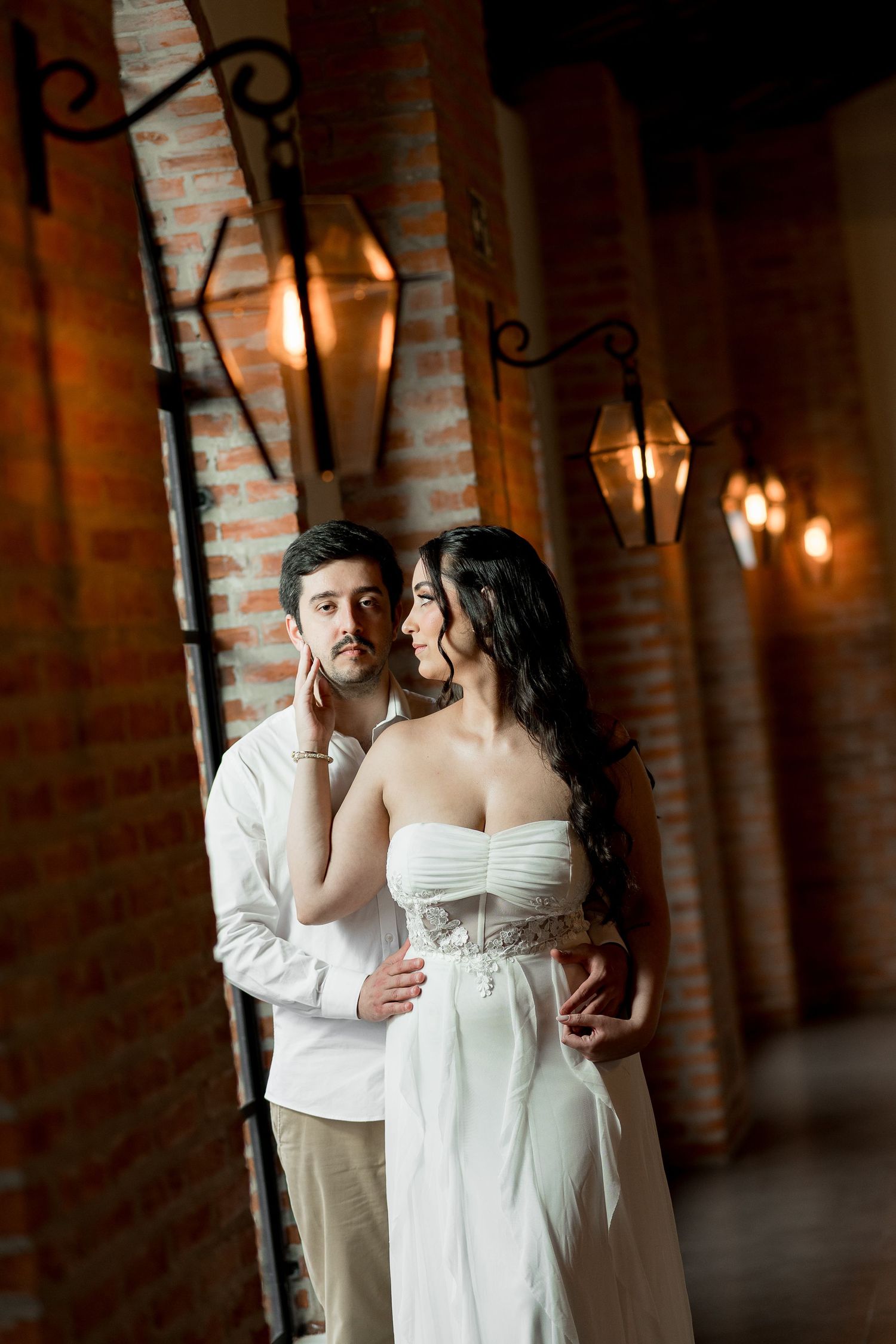 Romantic couple embracing near brick wall with vintage lanterns, woman in white strapless gown, man in white shirt.