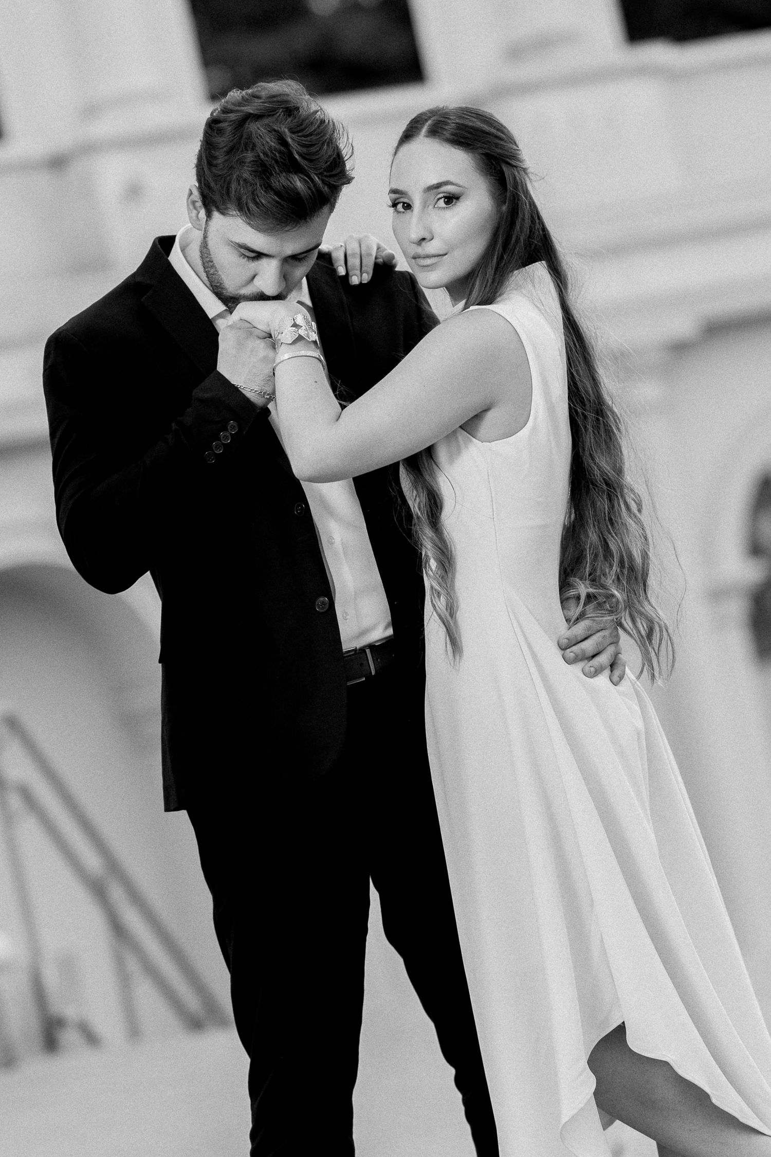 Black and white wedding photo of groom kissing bride's hand while she gazes at camera in elegant white dress.
