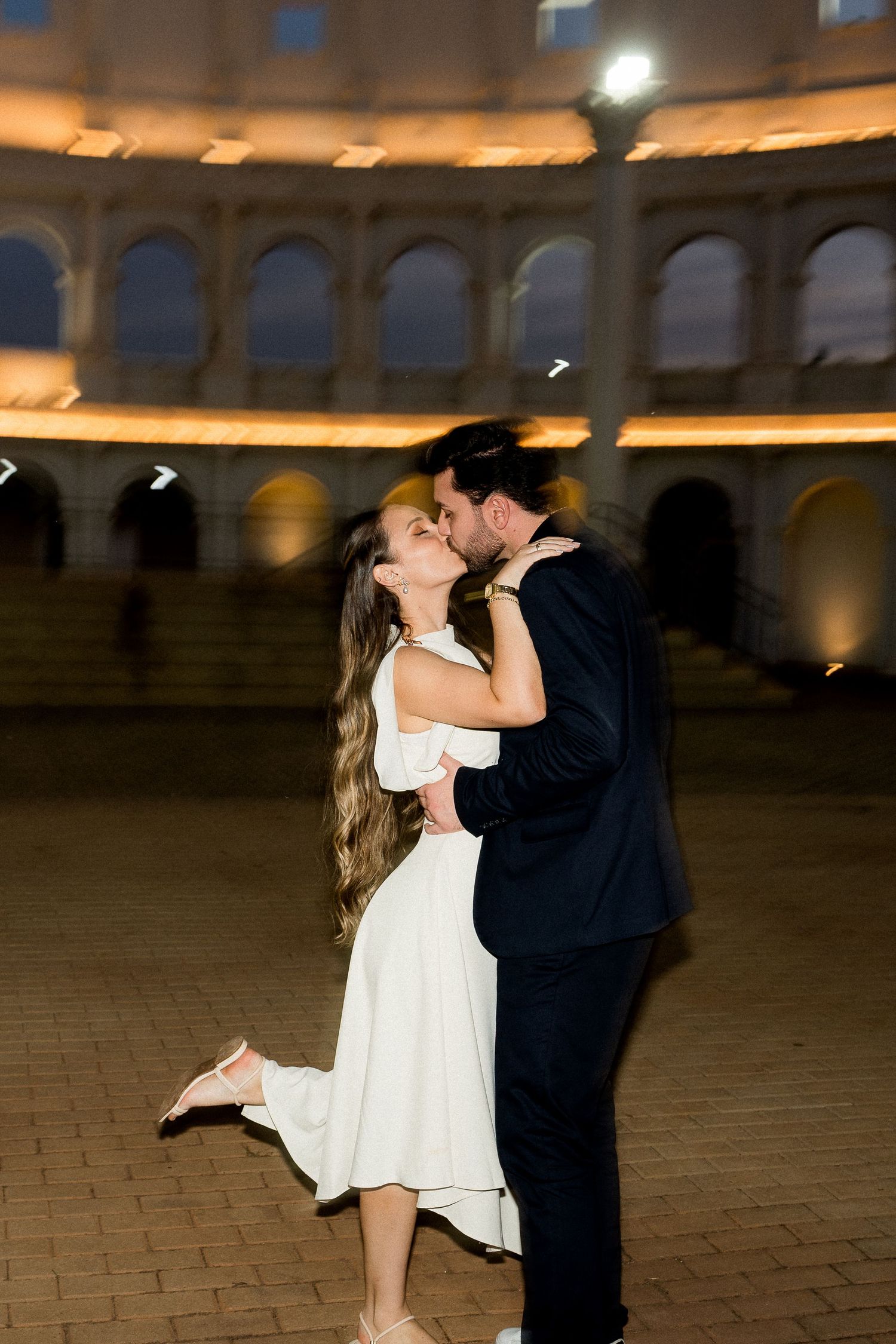 Couple sharing a romantic kiss at night in front of the illuminated Roman Colosseum, woman in white dress.