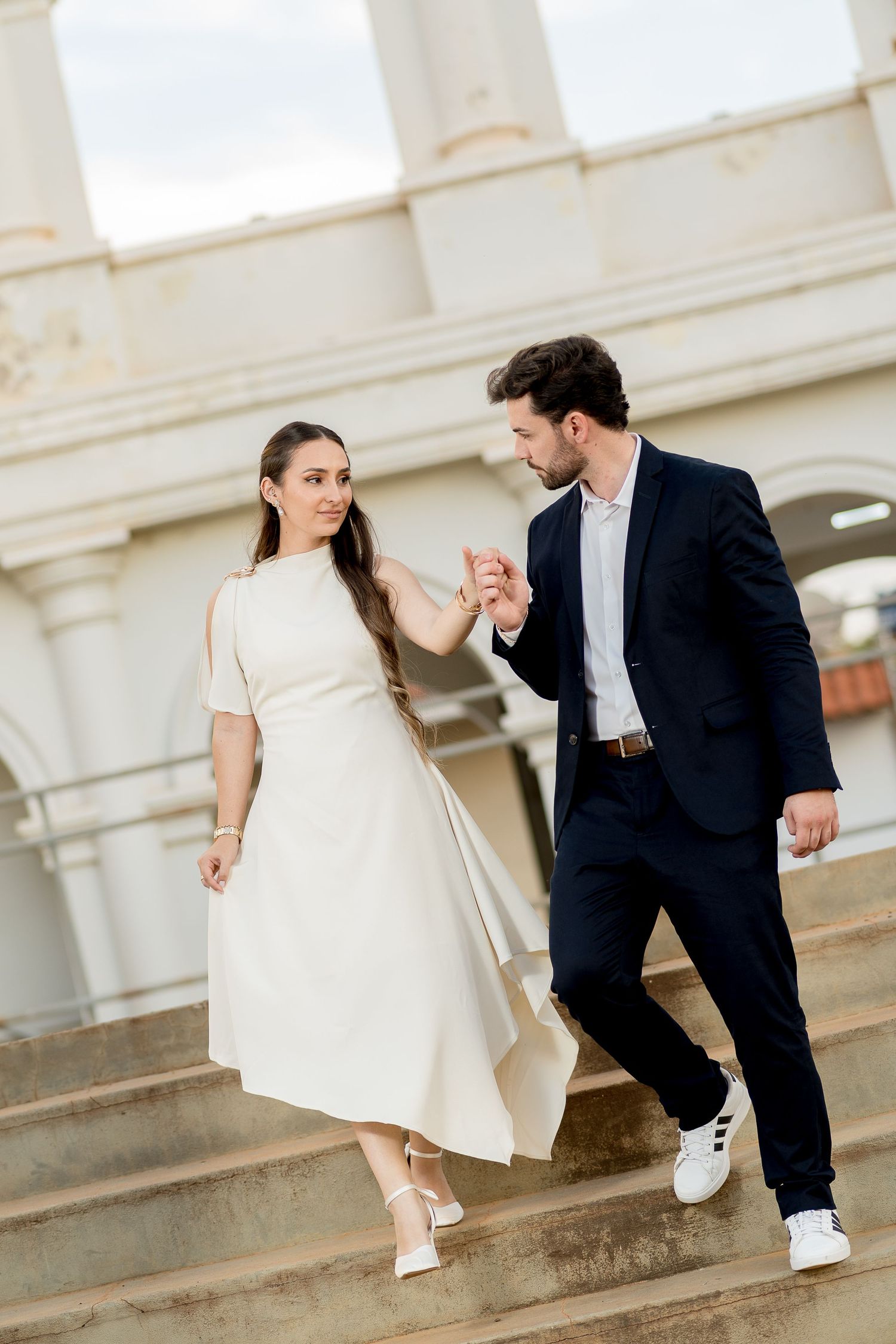 Elegant couple holding hands walking down outdoor steps, woman in white midi dress, man in navy suit.