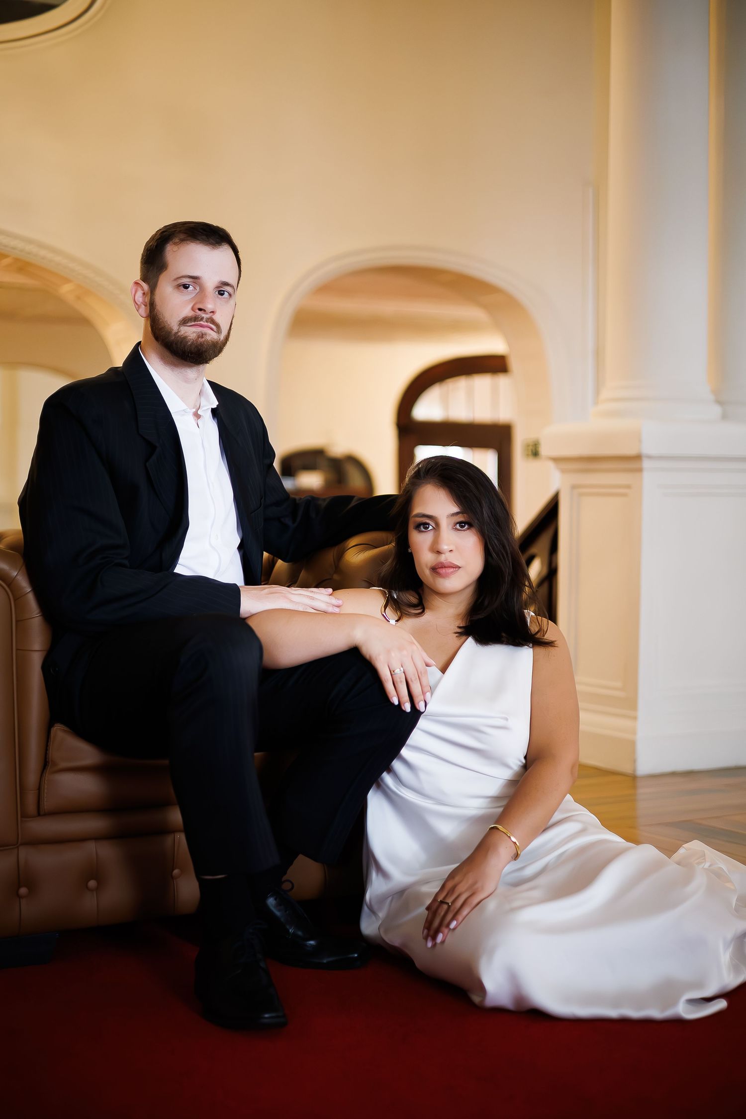 Elegant couple in formal attire posing in a grand interior, man in black suit seated, woman in white dress on floor.