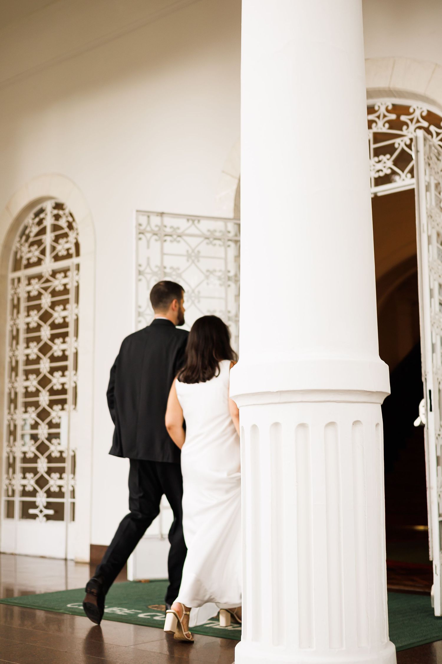 A bride in a white gown and groom in a black suit walk toward an elegant white building with ornate arched windows.