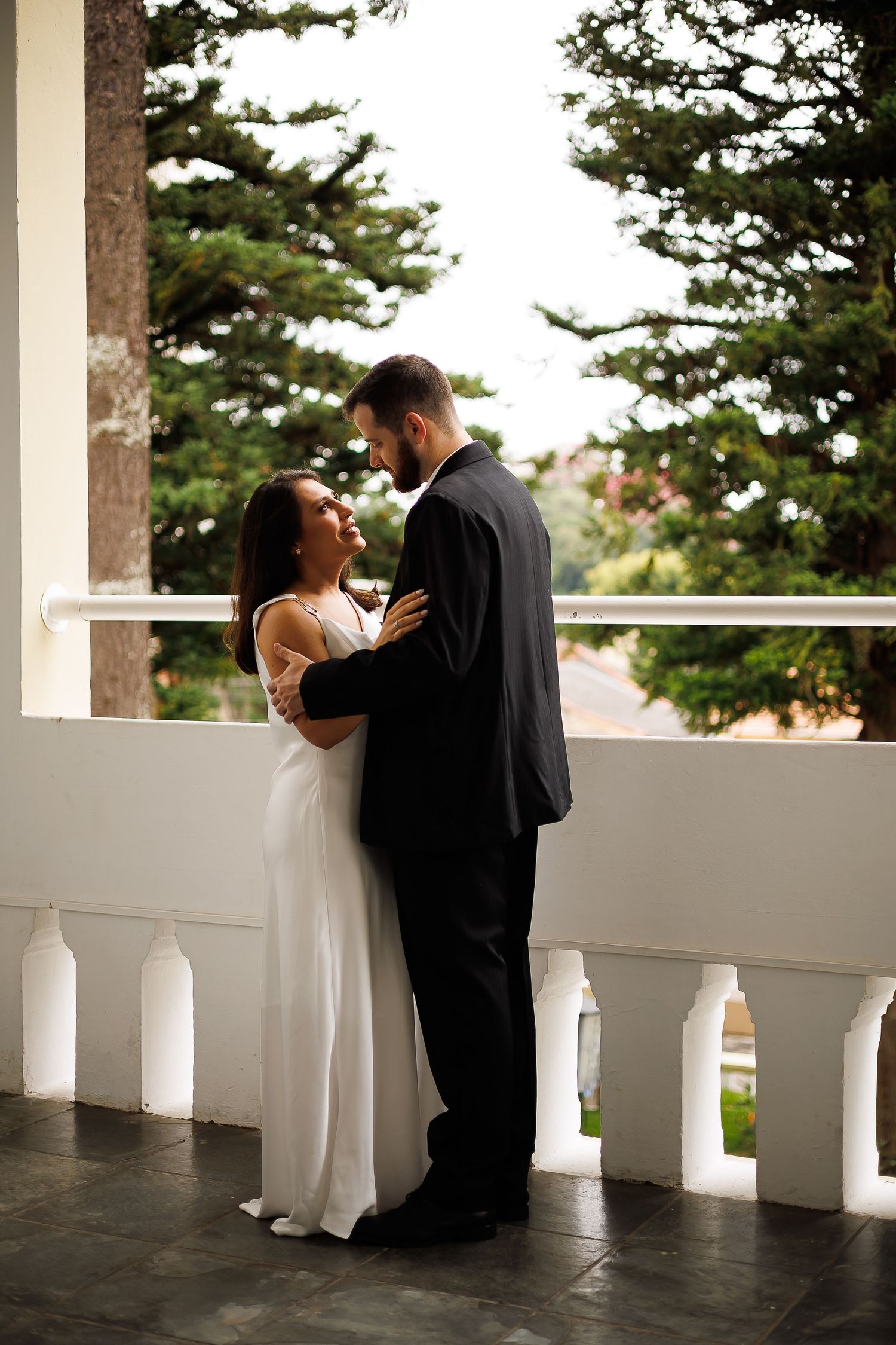 Bride in white gown and groom in black suit embrace on a balcony with lush green trees in the background.