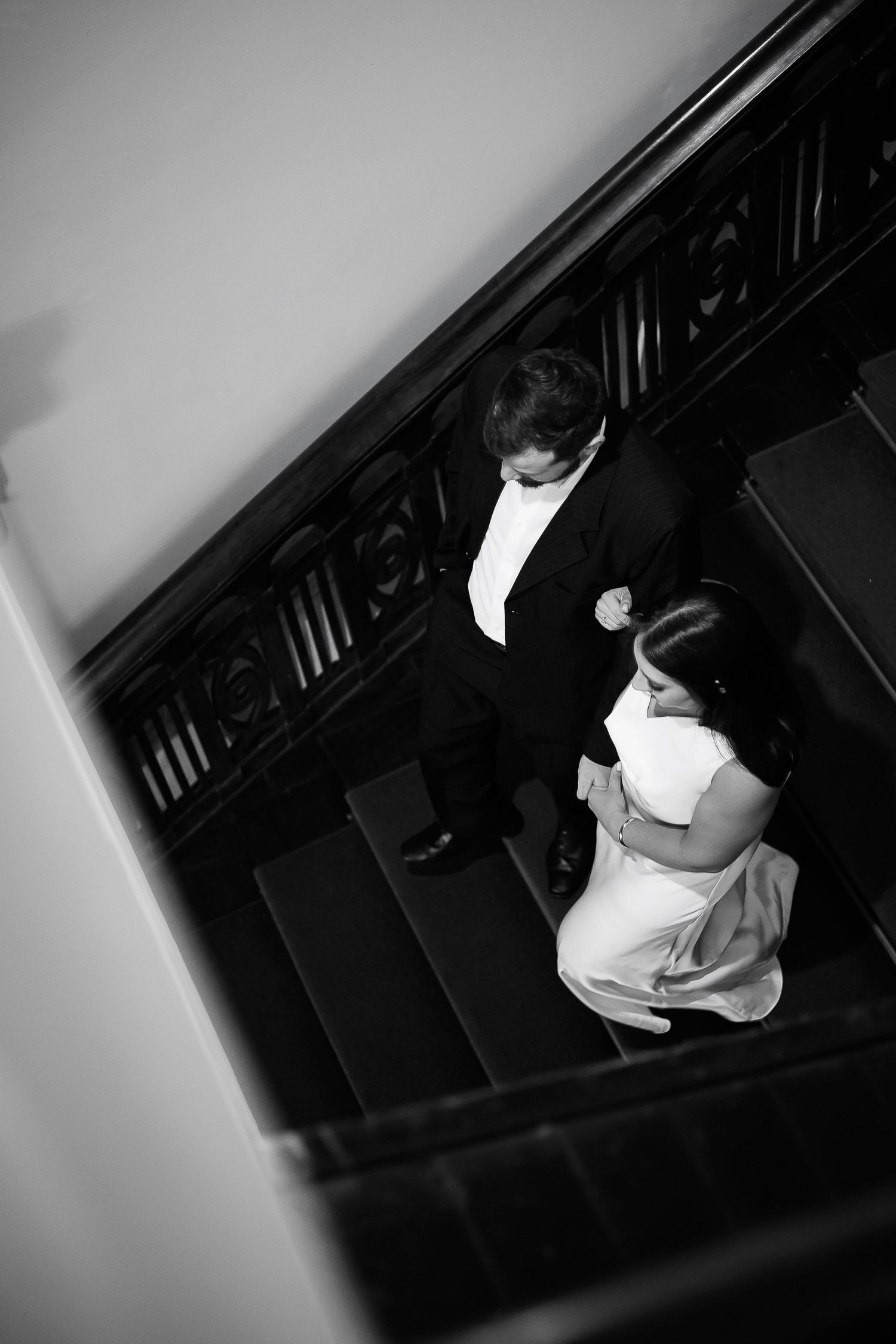 Black and white aerial view of a bride and groom in formal attire descending an elegant staircase.