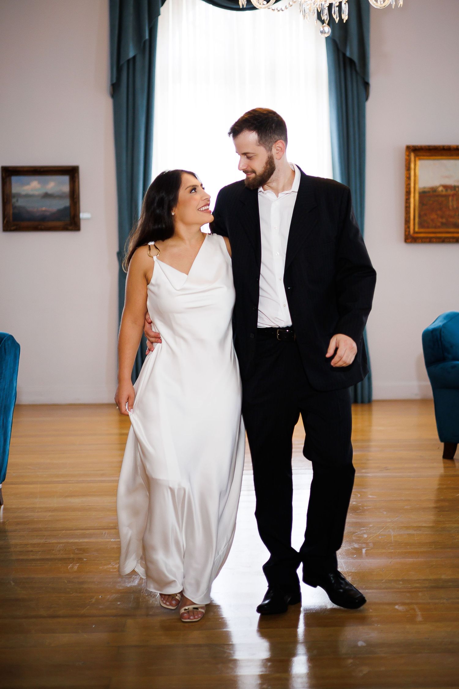 Bride in white gown and groom in black suit smiling at each other while walking in an elegant room.