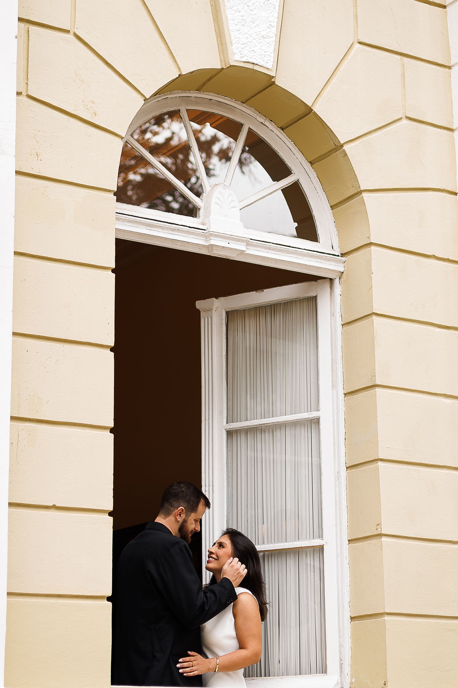 Bride and groom share a romantic moment in an elegant arched doorway of a cream-colored building.