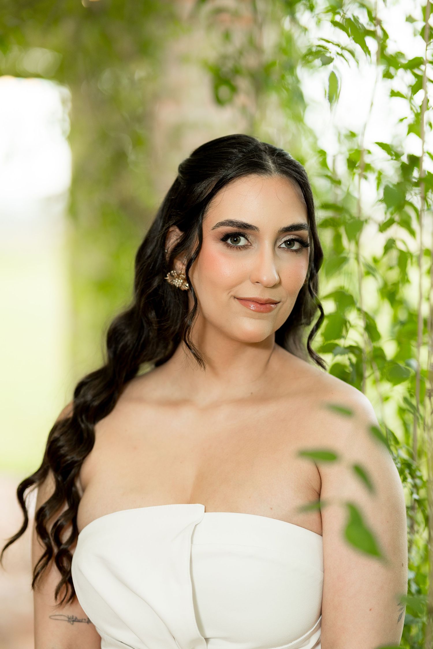 Bride in white strapless gown with long wavy dark hair and gold earrings posing among green leaves outdoors.