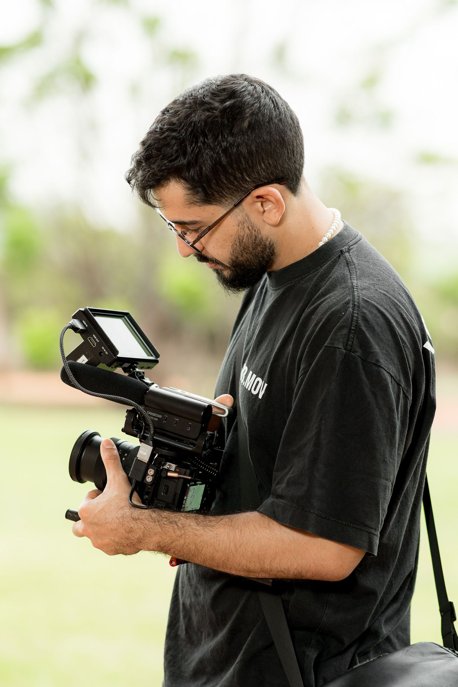 A bearded man in a black t-shirt holds a professional video camera outdoors, reviewing footage on a monitor.