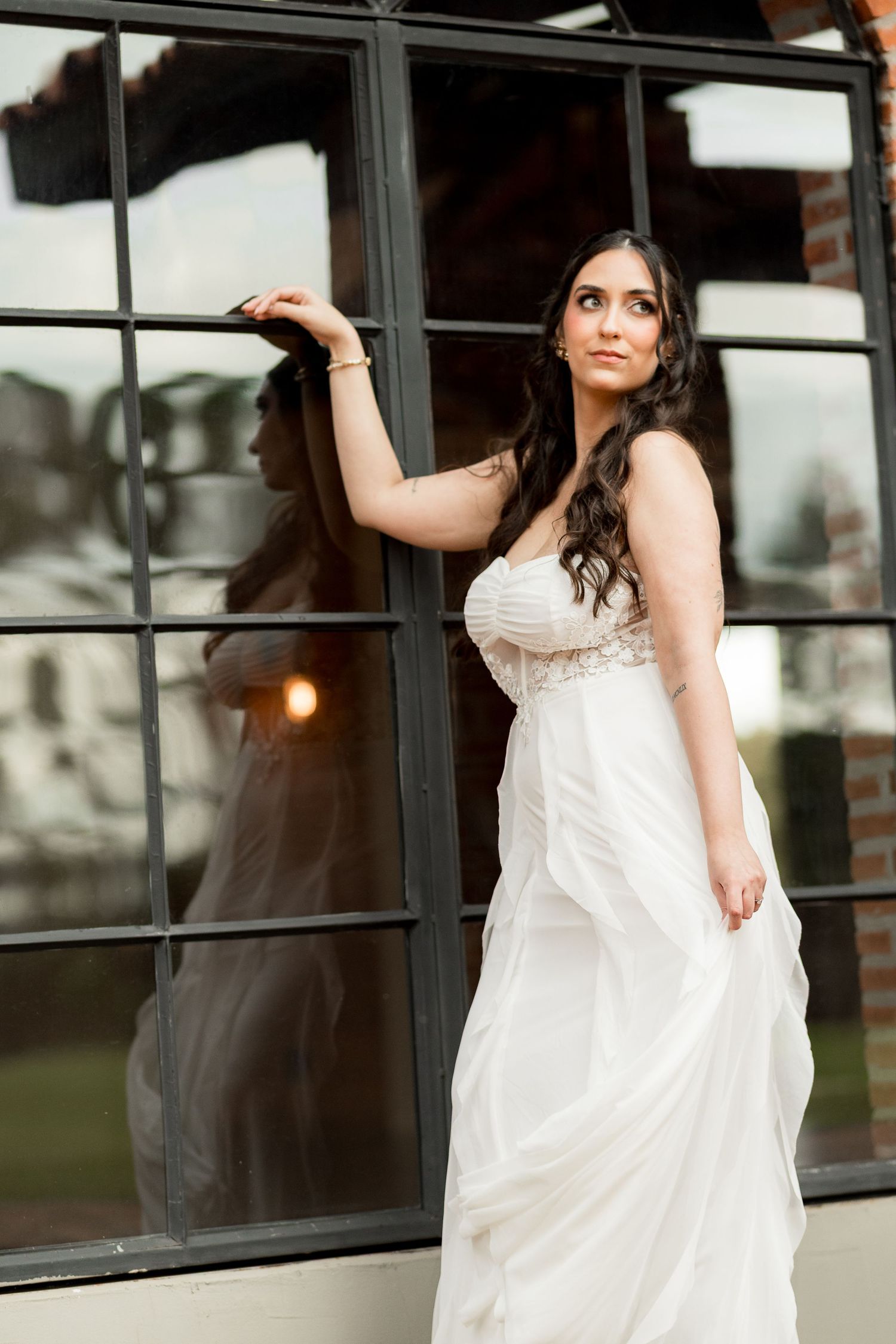 Bride in white flowing gown poses gracefully by large black-framed windows, her reflection visible in the glass.