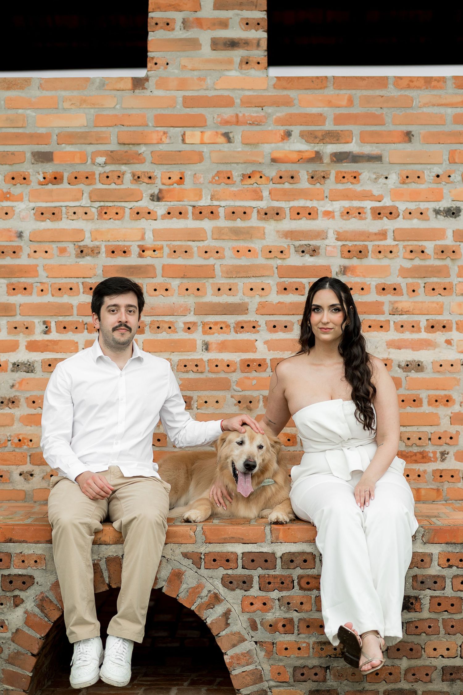 Couple in white and beige outfits sitting with a golden retriever dog against a rustic brick wall backdrop.
