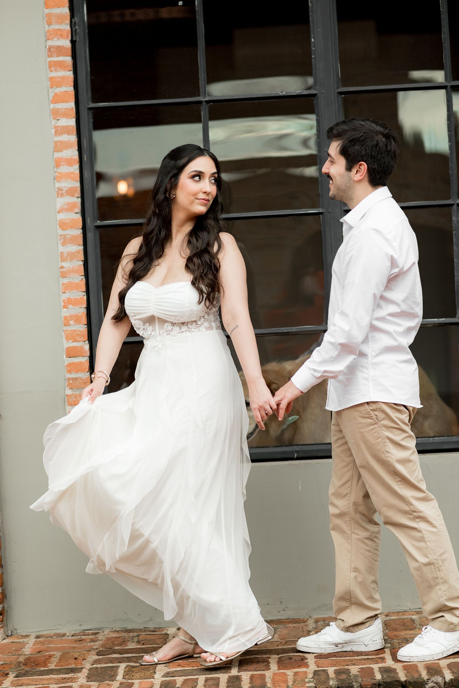 Couple in white attire holding hands and smiling at each other outdoors near a brick wall and large window.