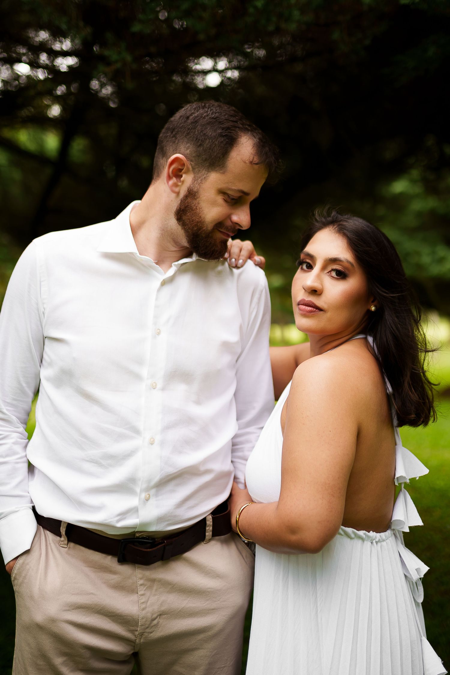 A couple in white attire poses romantically outdoors, the man kissing the woman's hand in a lush green setting.