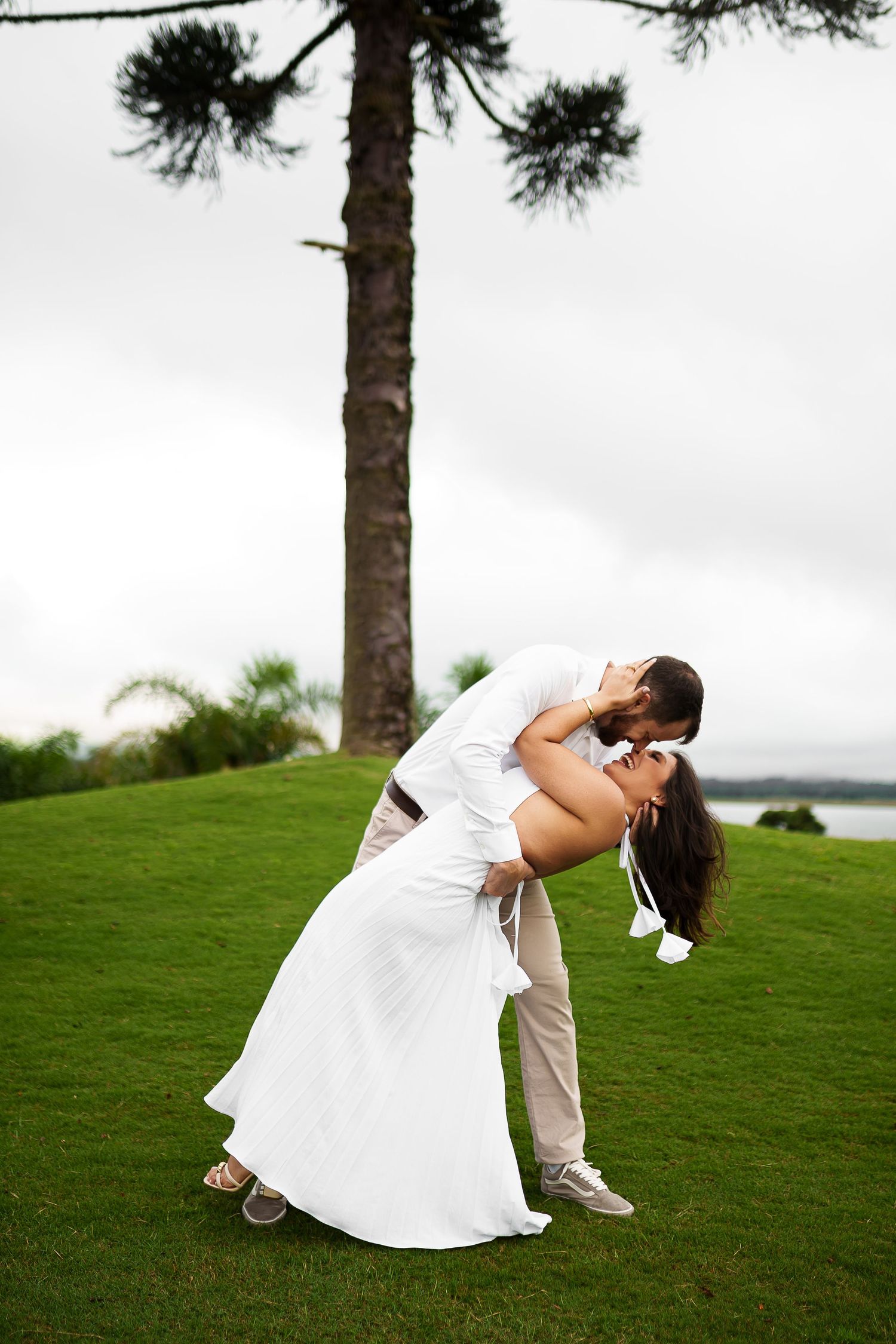 Couple sharing a romantic dip kiss on lush green lawn with tall pine tree backdrop on cloudy day.