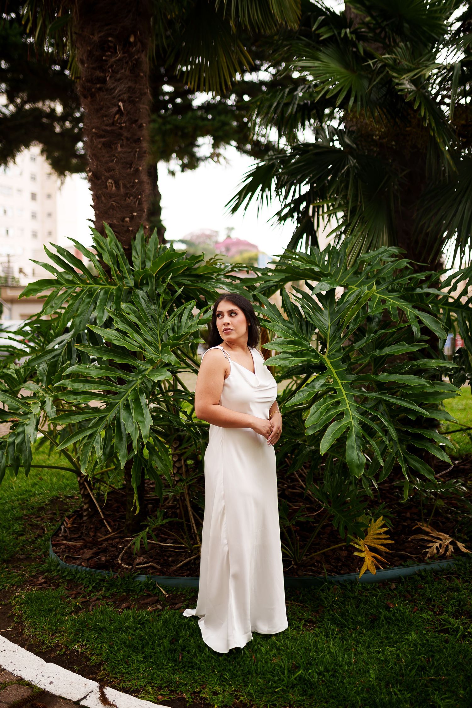 Woman in white satin gown standing among tropical palm trees and monstera leaves in lush green garden.