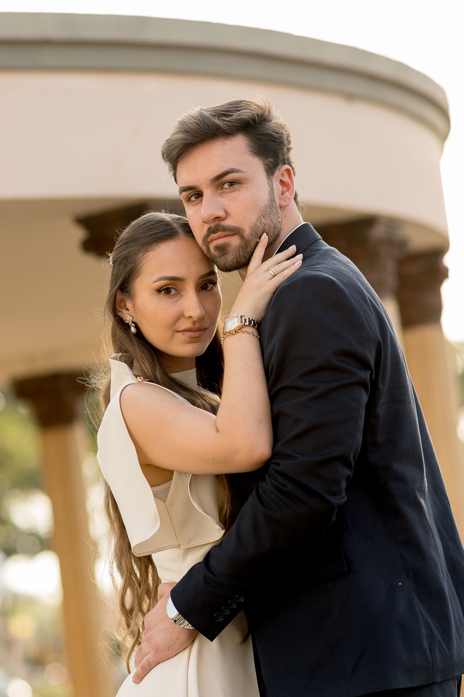 Elegant couple embracing outdoors, woman in white dress and man in dark suit, posing near classical columns.
