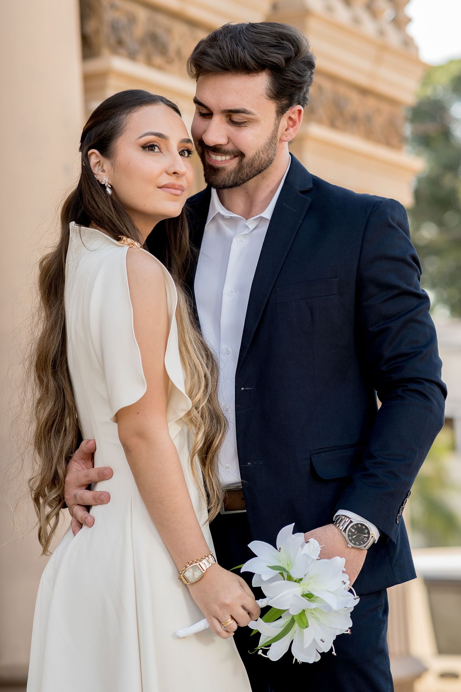 Elegant couple in wedding attire, bride in white dress holding white lilies, groom in dark suit smiling outdoors.