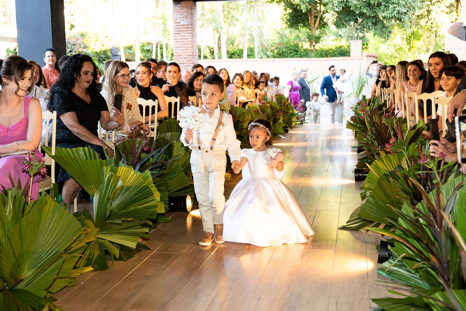 Two young children dressed in white walk down a floral-lined wedding aisle as flower girl and ring bearer.