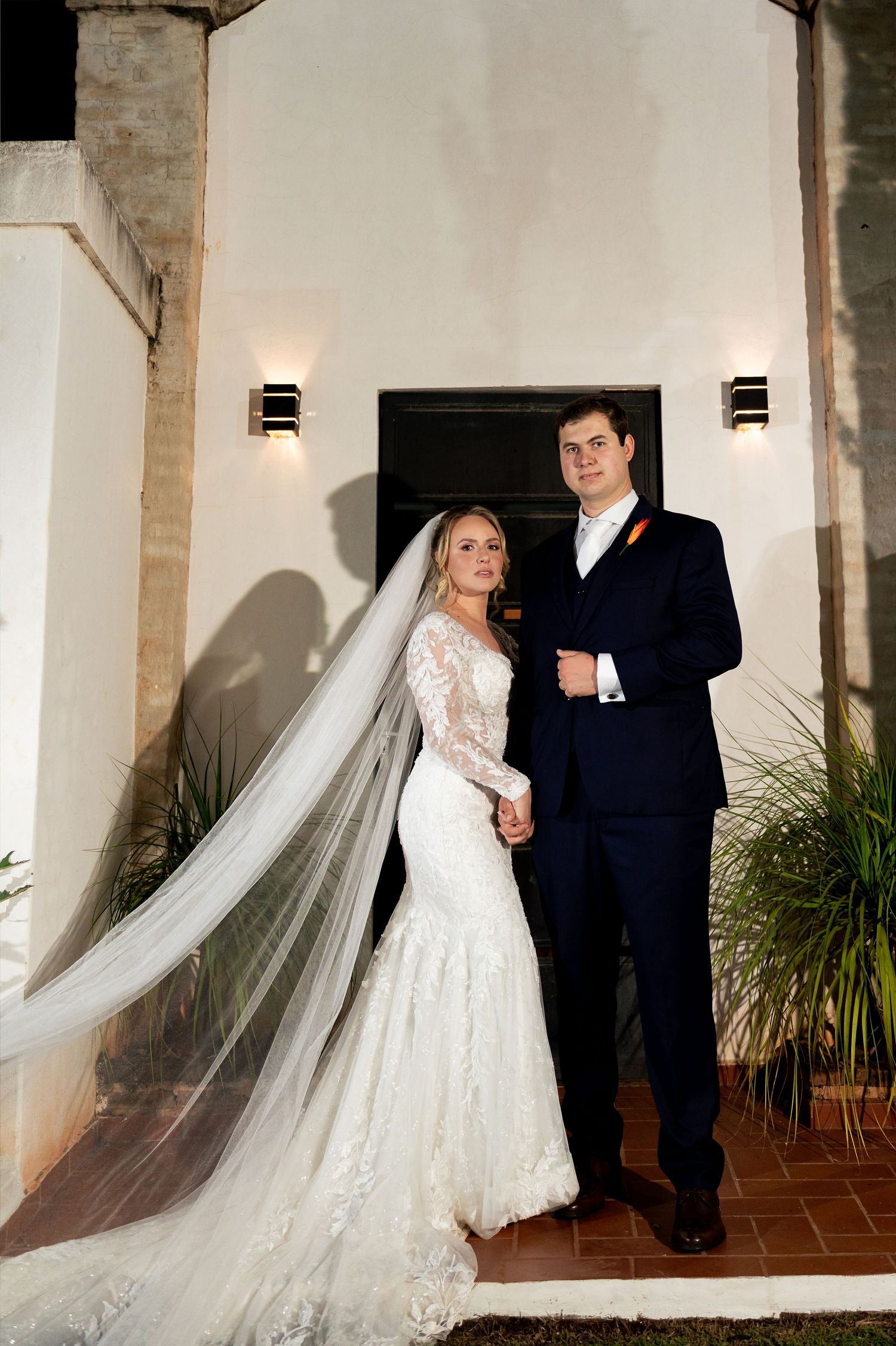 Bride in lace mermaid gown with flowing veil and groom in navy suit hold hands outside elegant venue at night.