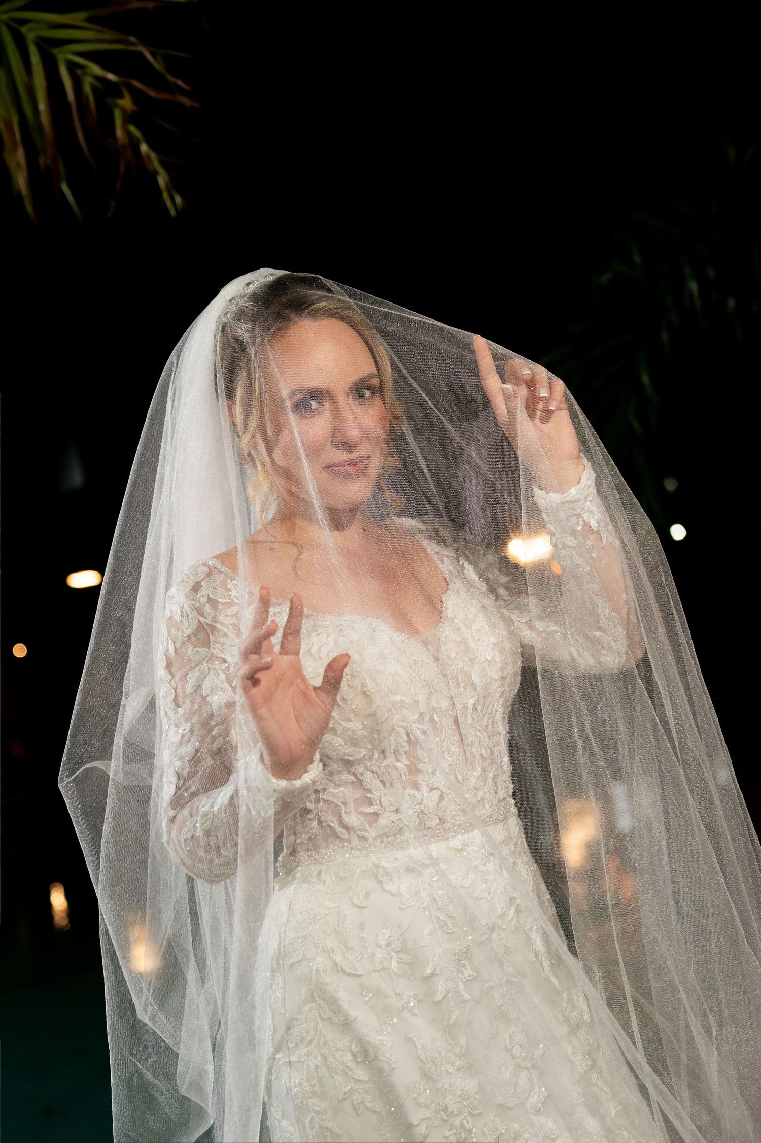 Bride in lace long-sleeve wedding dress lifts veil with both hands making peace signs at night.