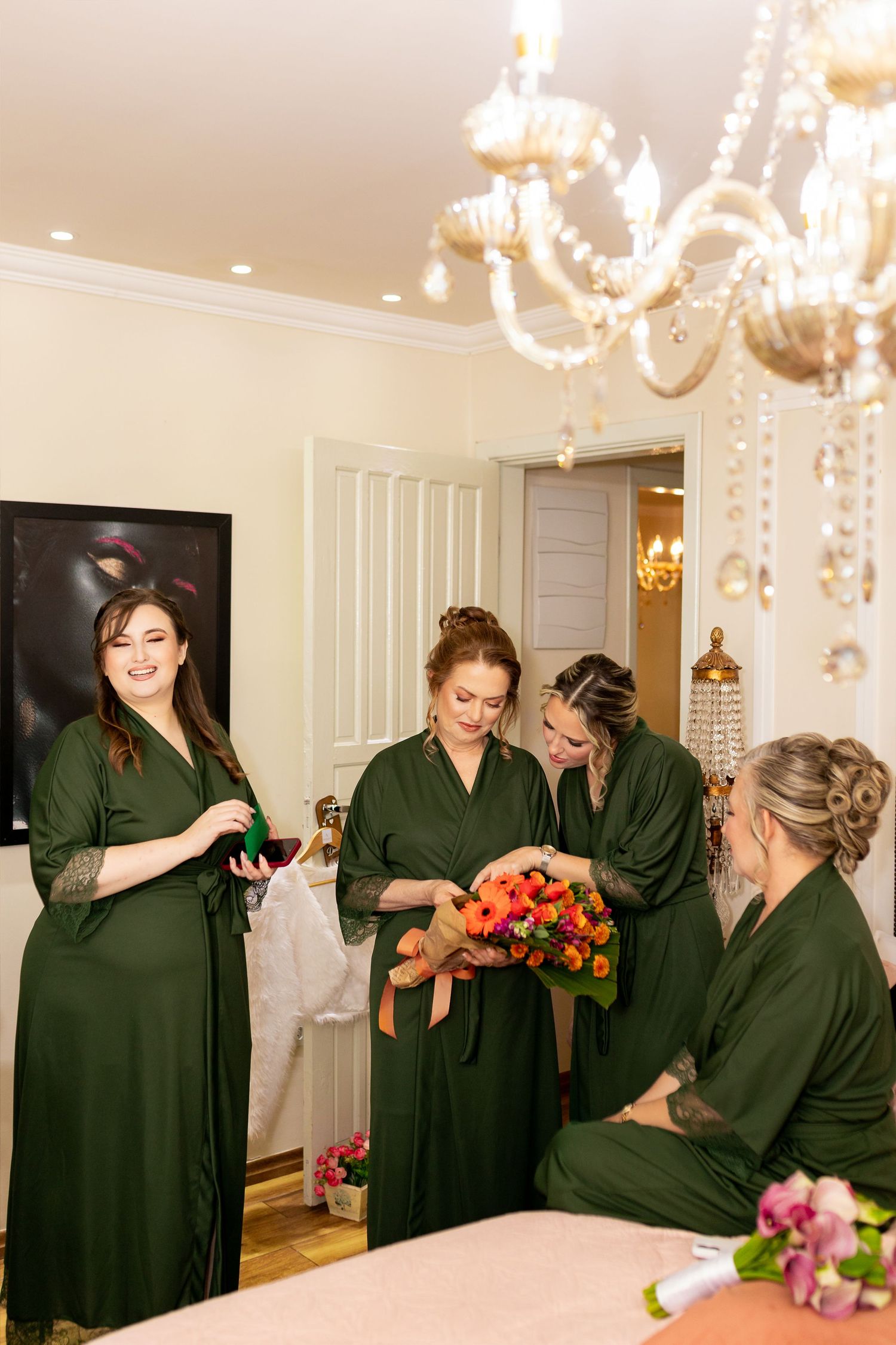 Bridesmaids in green robes admire a colorful bridal bouquet in an elegant room with a crystal chandelier.