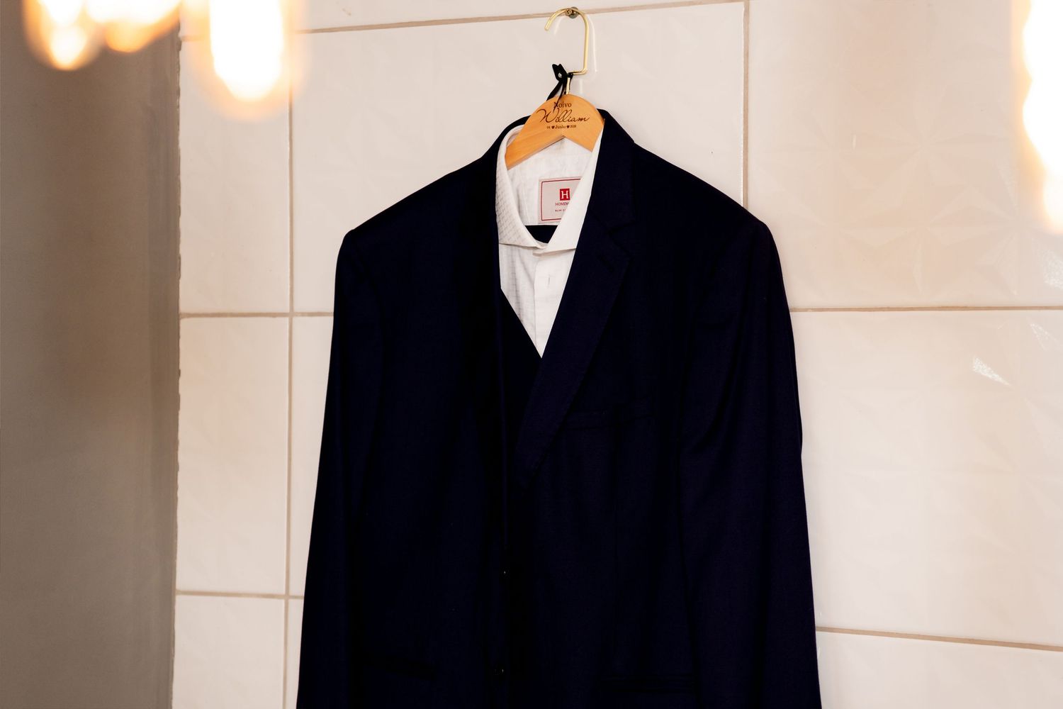 A dark navy suit jacket hanging on a wooden hanger against a white tiled wall with soft warm lighting.