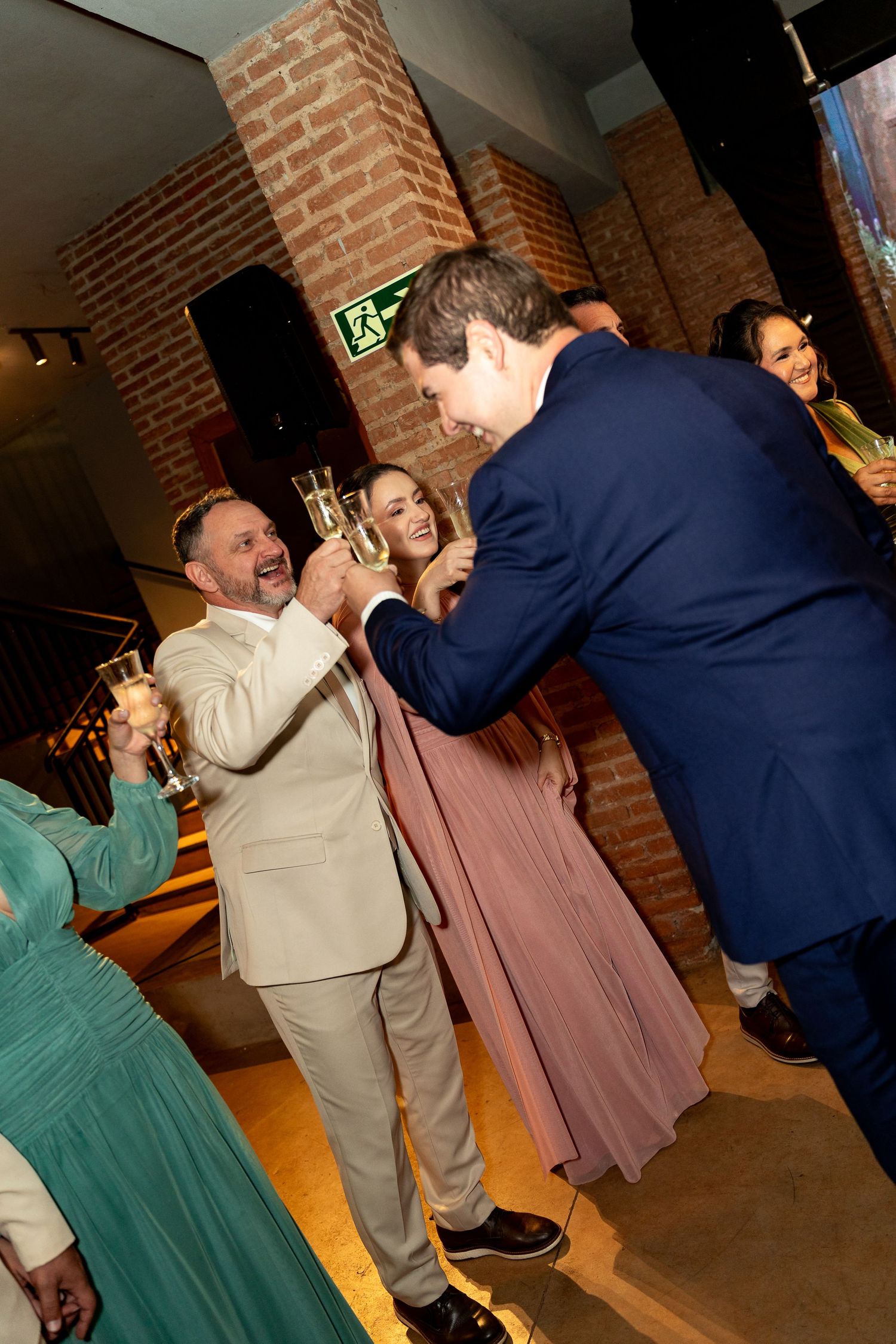 Guests toasting with champagne glasses at an elegant indoor event, wearing formal attire near a brick wall.