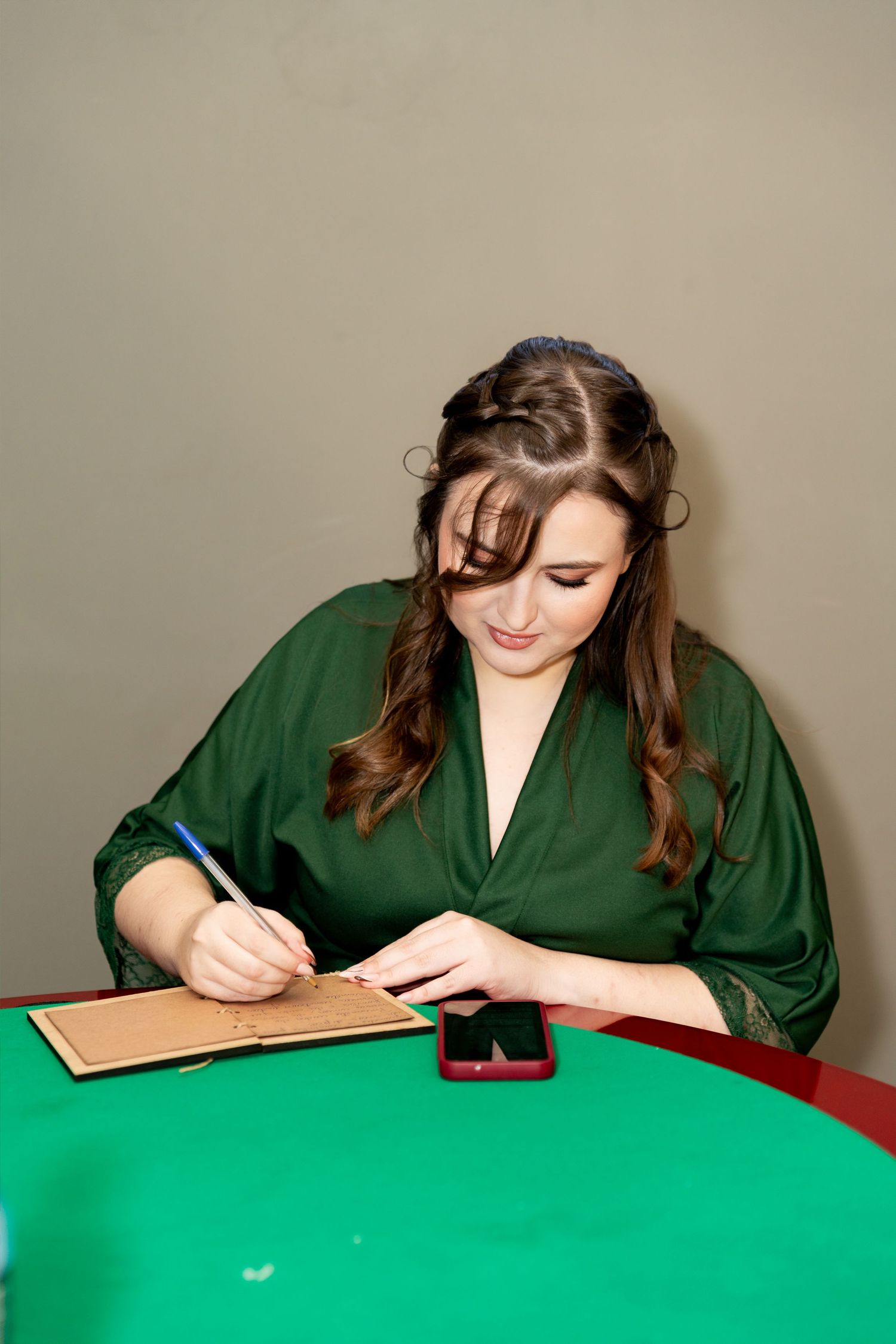 Young woman in green robe writing on clipboard at green table with smartphone nearby.