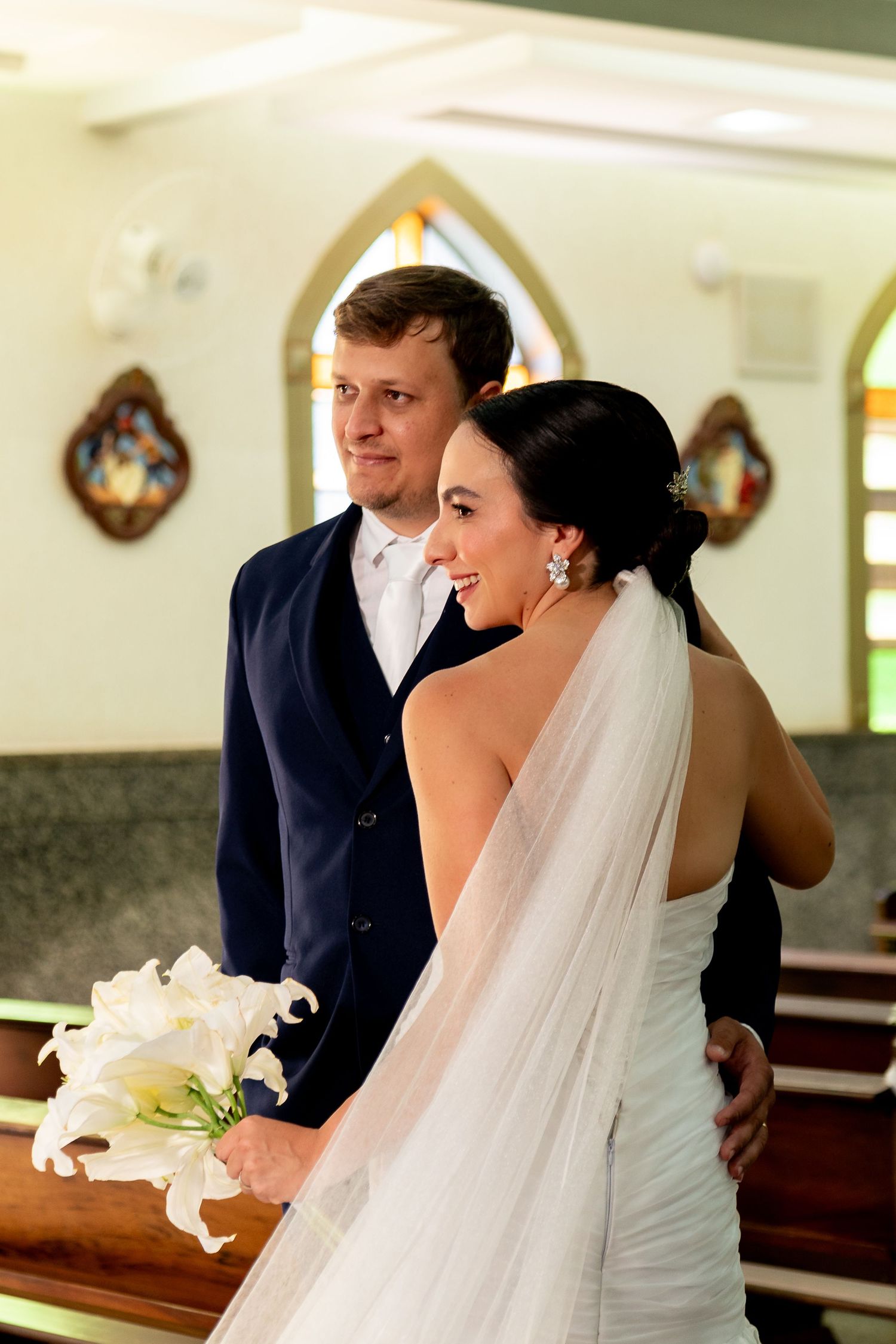 Bride in white gown holding calla lilies smiles at groom in navy suit during wedding ceremony in church.