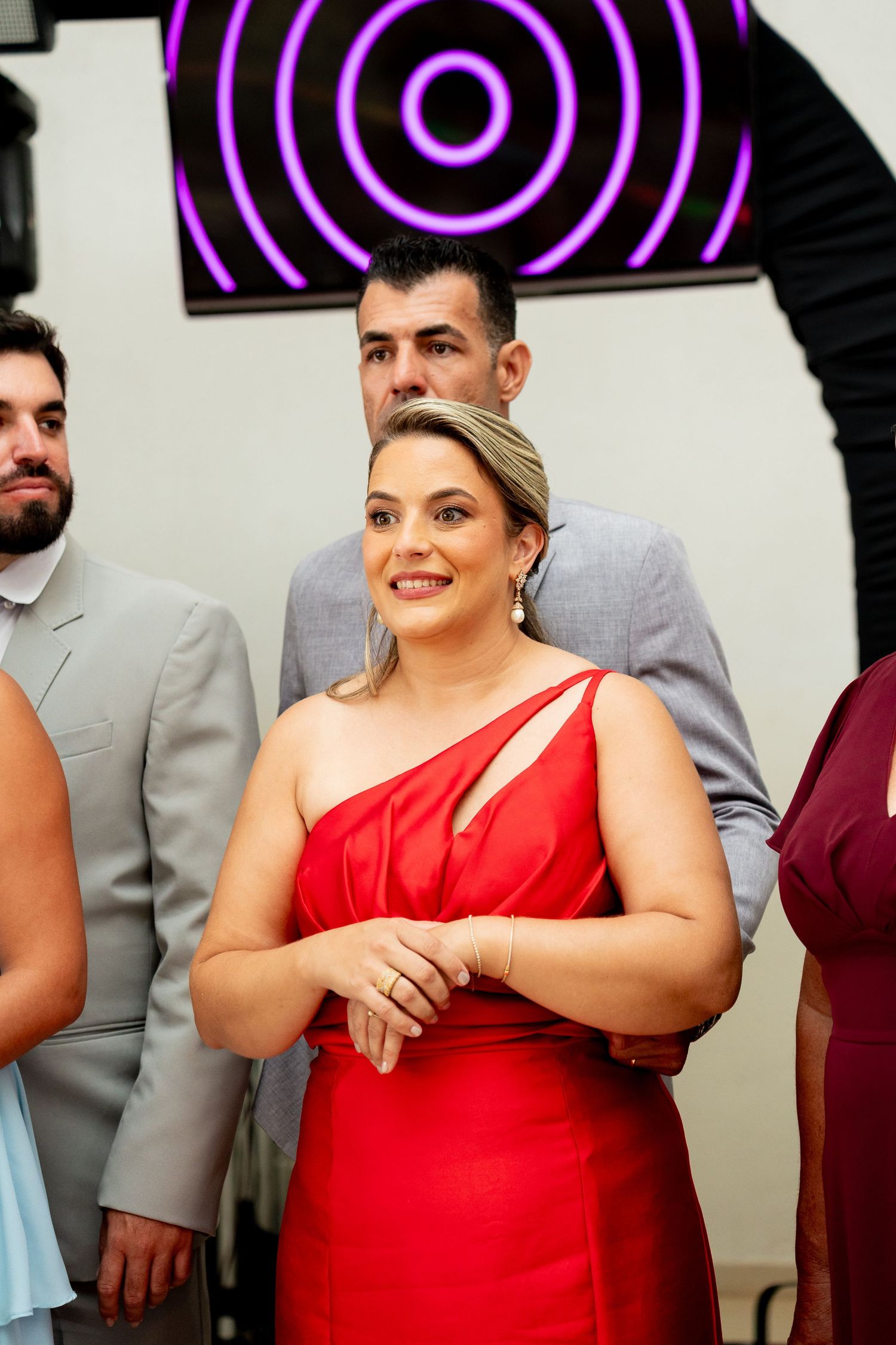 Woman in red one-shoulder dress smiling at formal event, with guests in suits and evening wear behind her.