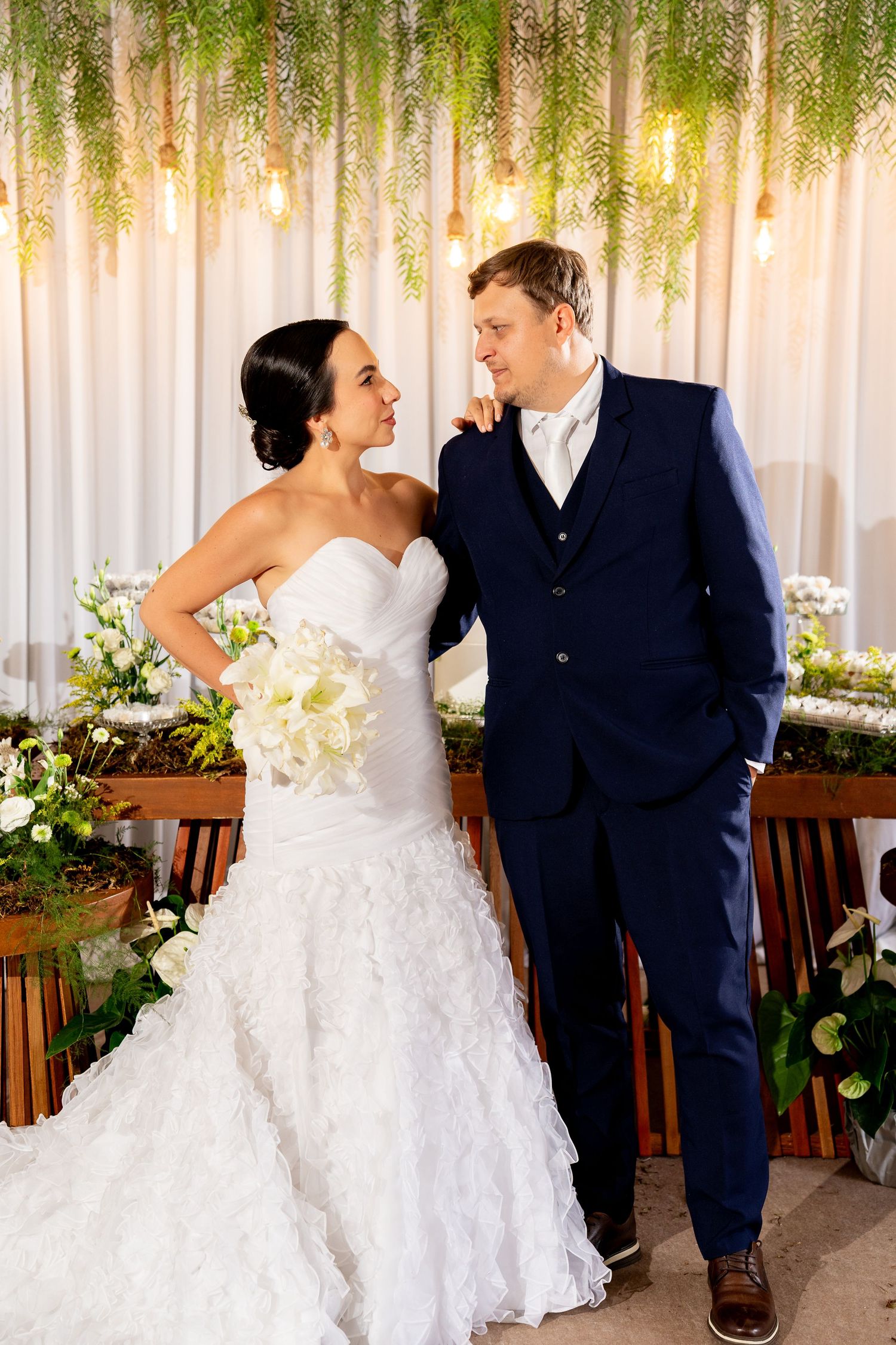 Bride in white mermaid gown holding bouquet stands with groom in navy suit before floral backdrop with hanging greenery.