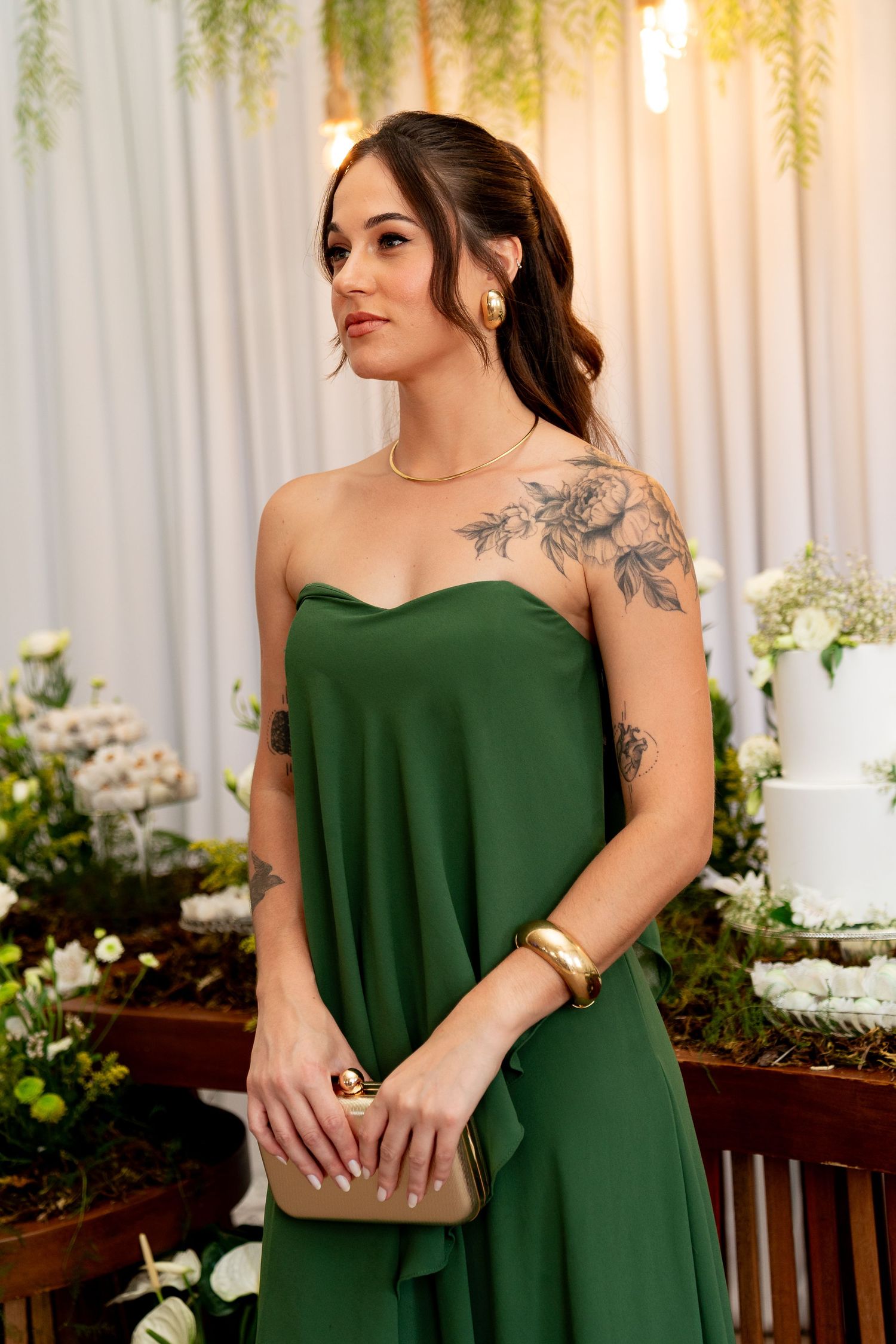 Woman in strapless green gown with floral tattoos holds gold clutch at elegant wedding reception.