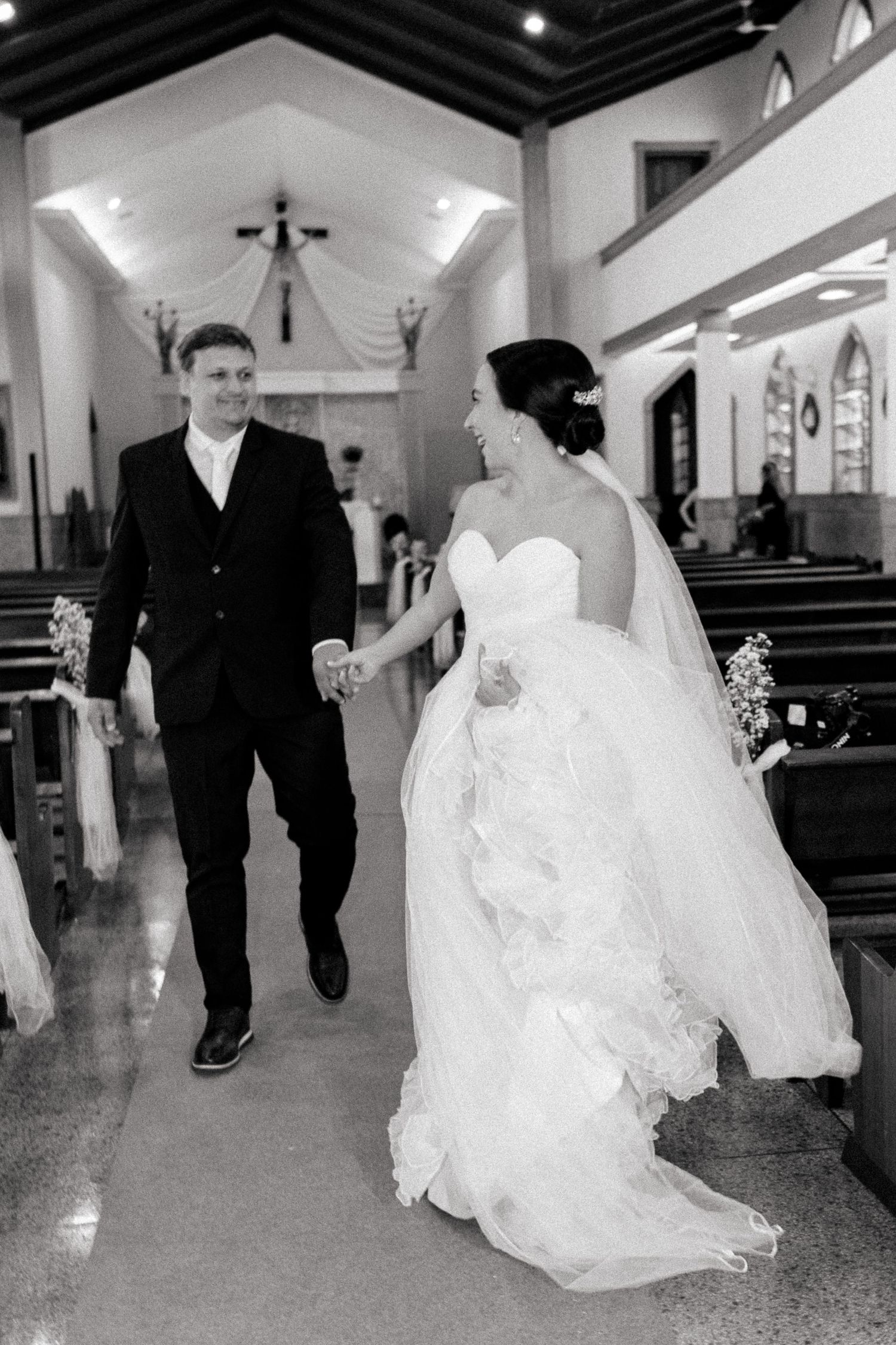 Black and white photo of a joyful bride and groom walking hand in hand down a church aisle after their wedding ceremony.