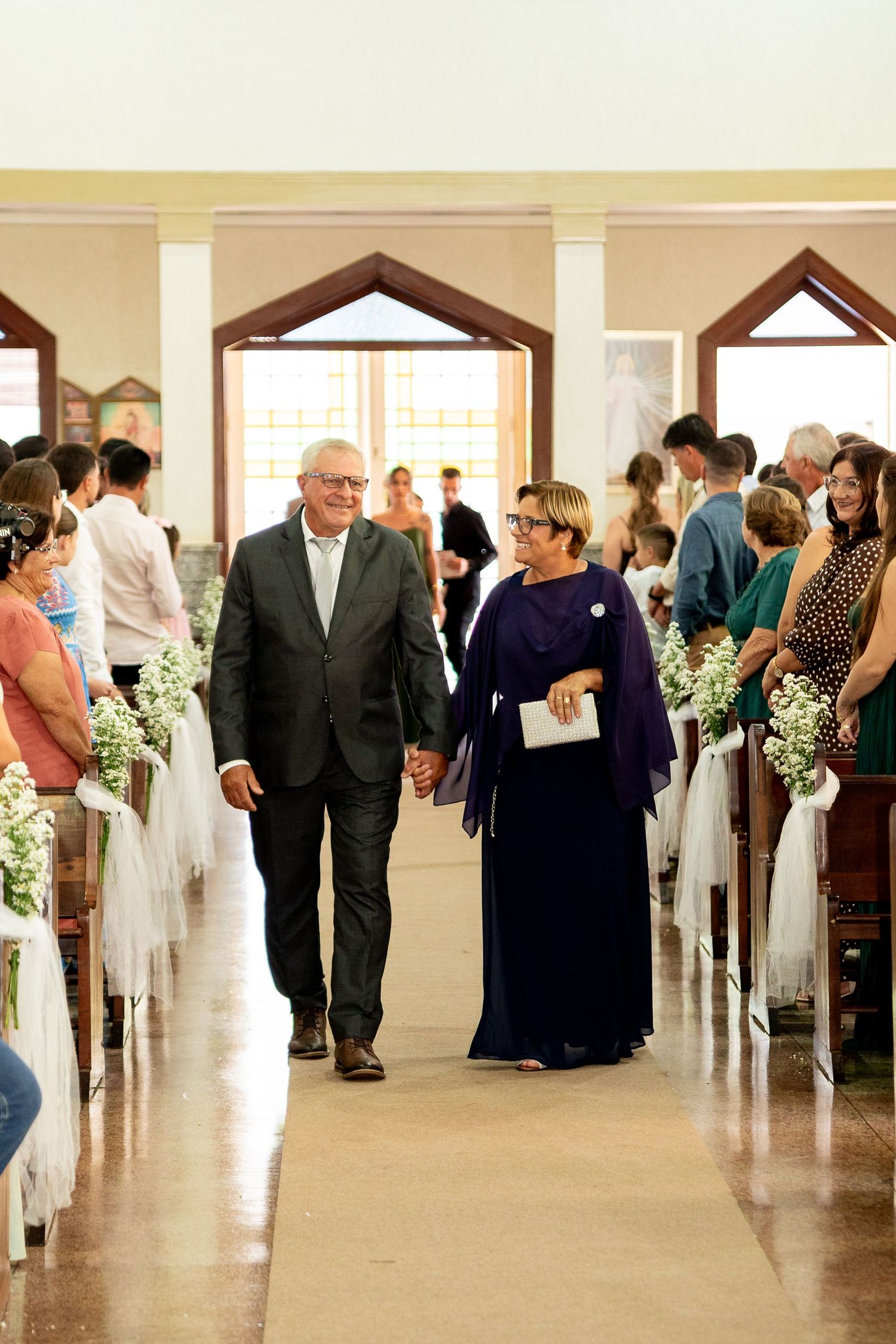 Elderly couple walks hand-in-hand down church aisle at wedding ceremony, guests seated on decorated pews.