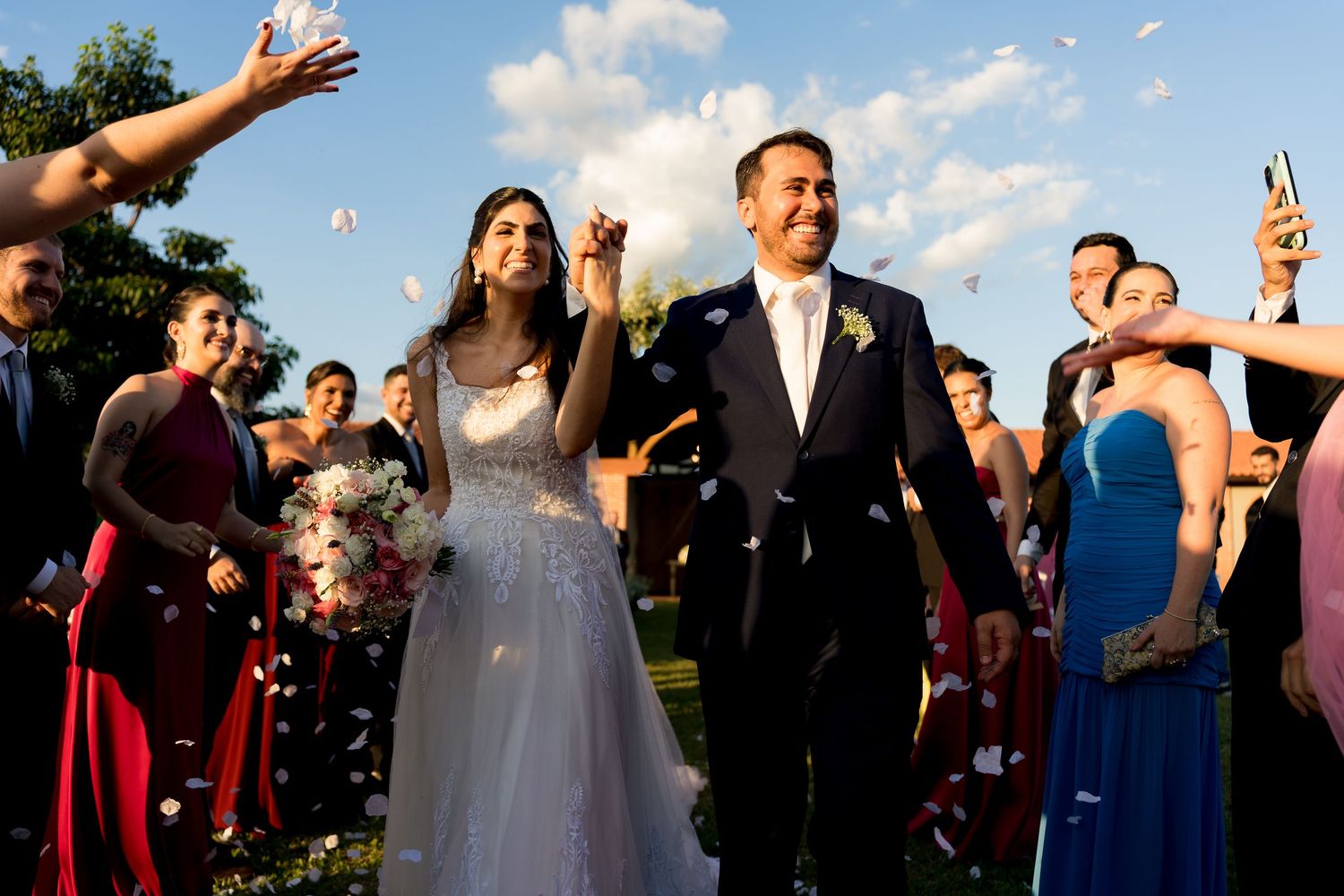 Bride and groom walk through confetti shower with wedding party cheering outdoors on a sunny day.
