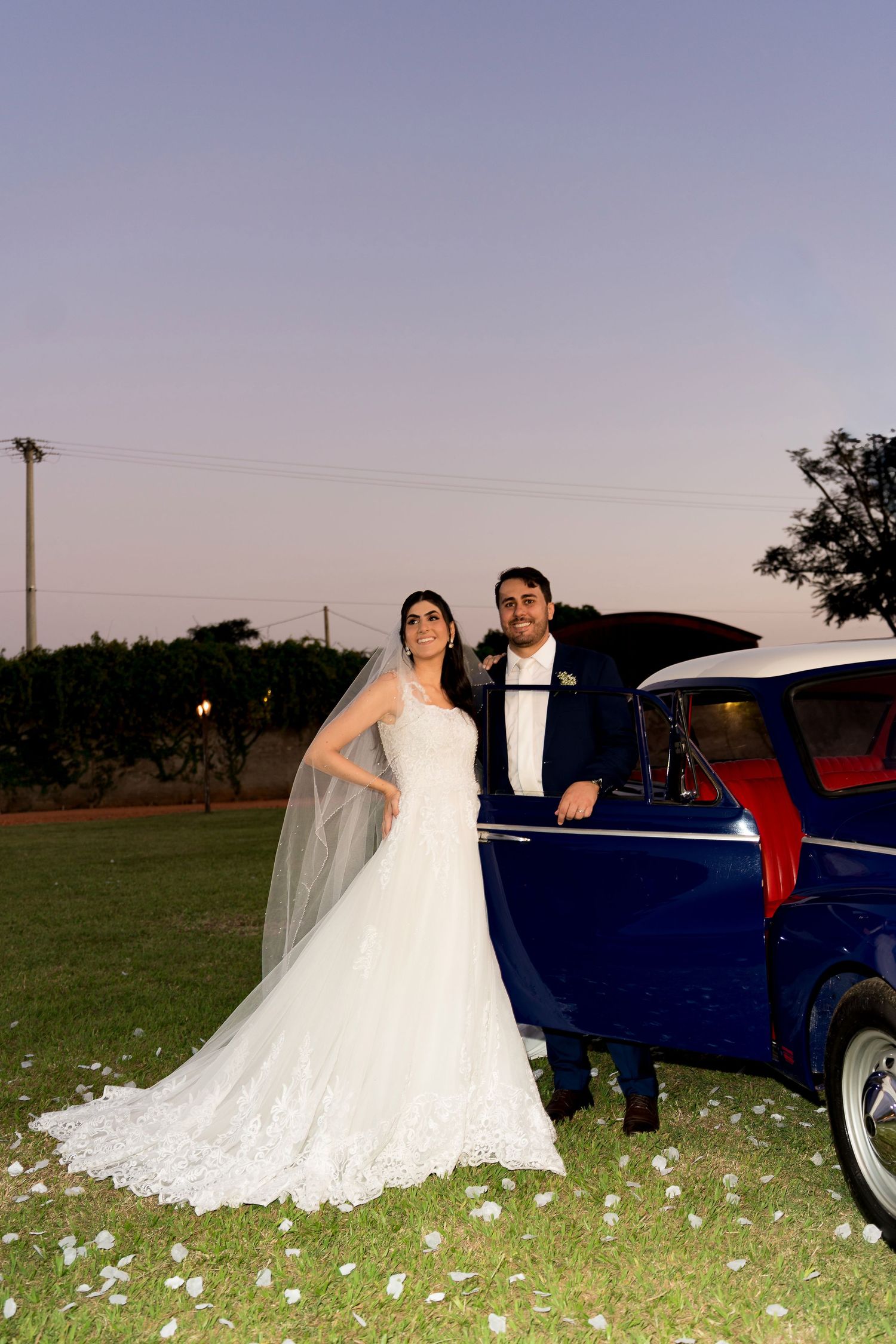 Bride in white lace gown with veil and groom in dark suit pose beside a classic blue car at dusk.