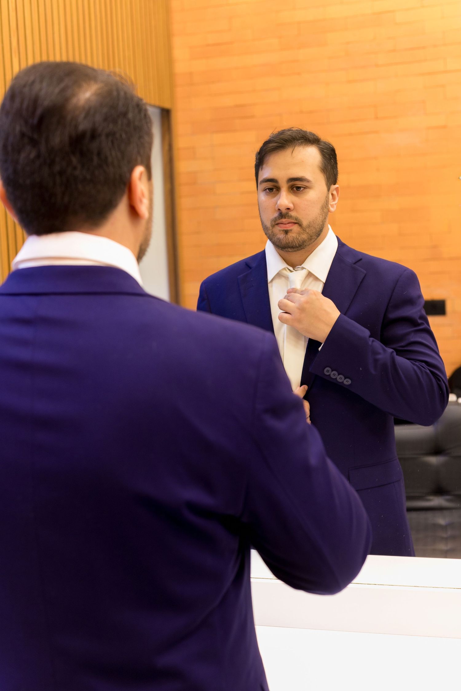 Man in blue suit adjusting his tie while looking at his reflection in a mirror.