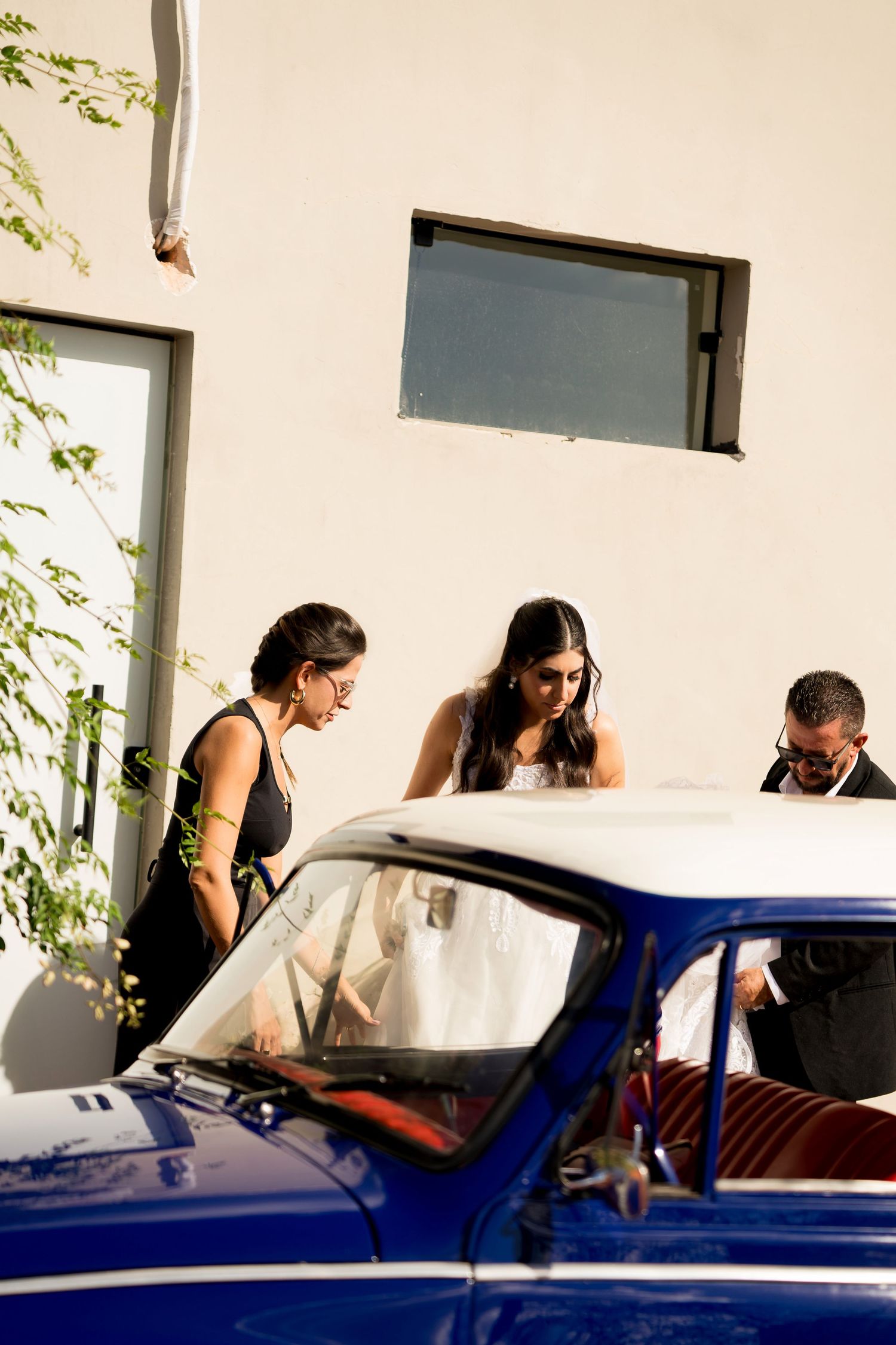 Bride in white gown with guests beside a vintage blue car outside a white building on a sunny wedding day.