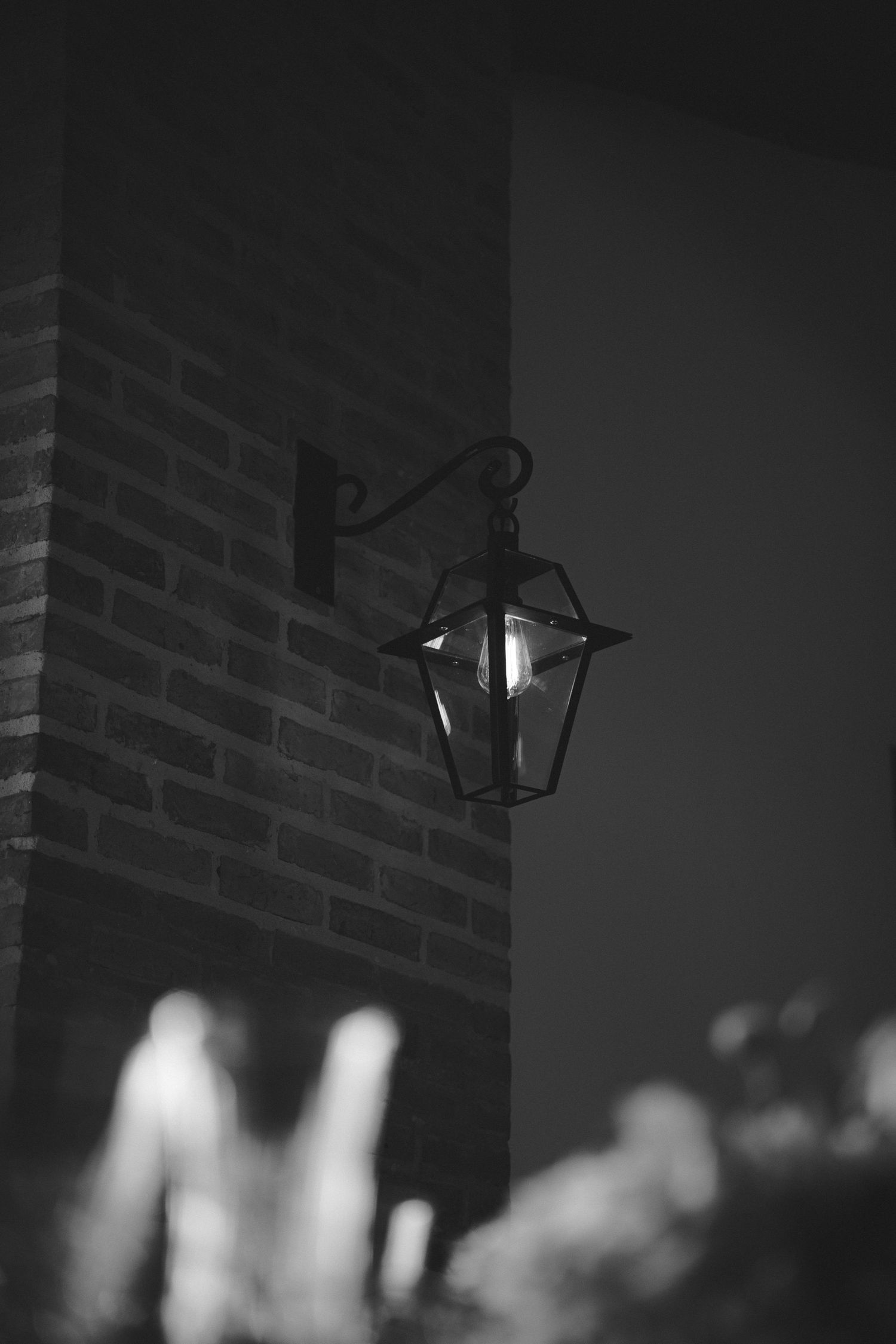 Black and white photo of a vintage wall-mounted lantern glowing on a brick building corner at night.