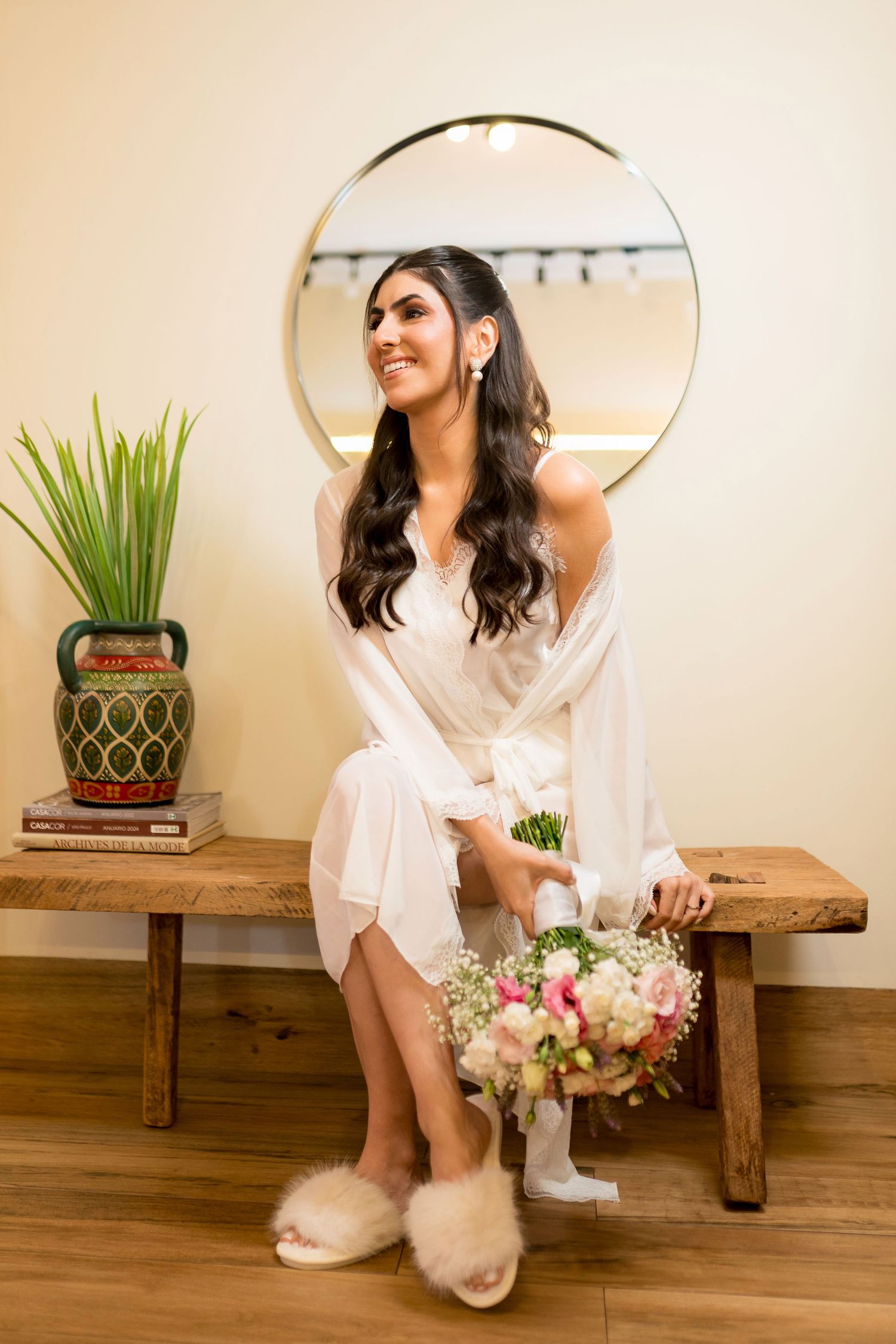 Smiling bride in white robe and fluffy slippers sits on wooden bench holding floral bouquet before a round mirror.